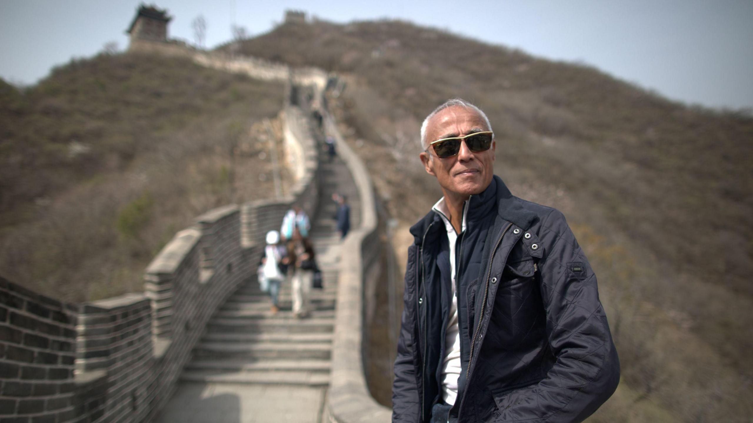 A recent photo of Andrew Ridgeley, sitting against the Great Wall of China. He has grey cropped hair, is wearing sunglasses, a navy jacket and white top.