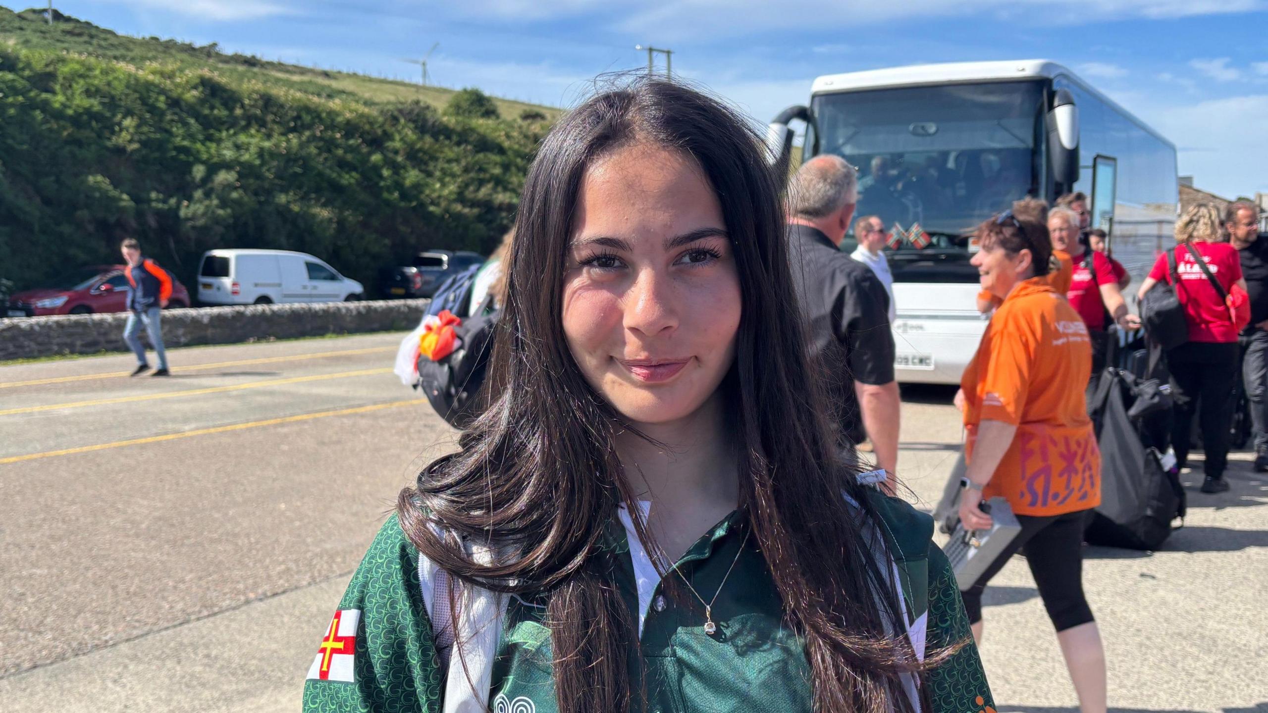 Alexia Nascimento is standing in front of a coach smiling into the camera. She is wearing Guernsey kit.