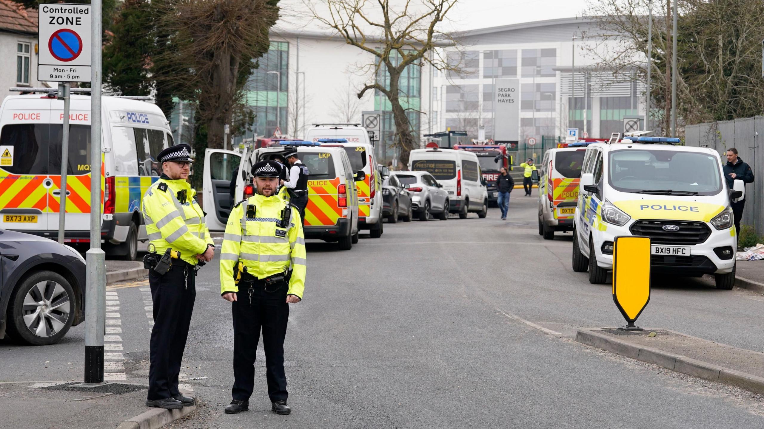 Uniformed police officers stand beside numerous police vans on a road near the scene