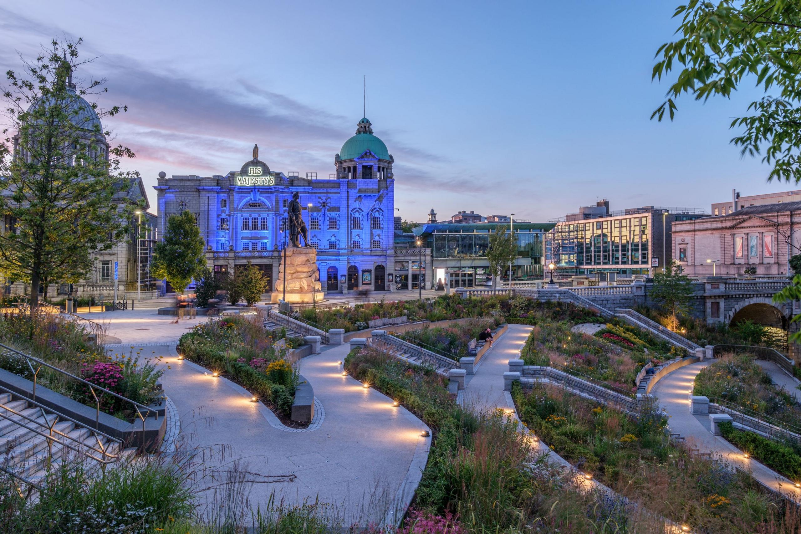 A view of Union Terrace Gardens looking to His Majesty's Theatre, taken in the evening as the sun goes down and the garden lighting comes on. Clean, light pathways run across the multi-level space, with established plants in between the walkways.