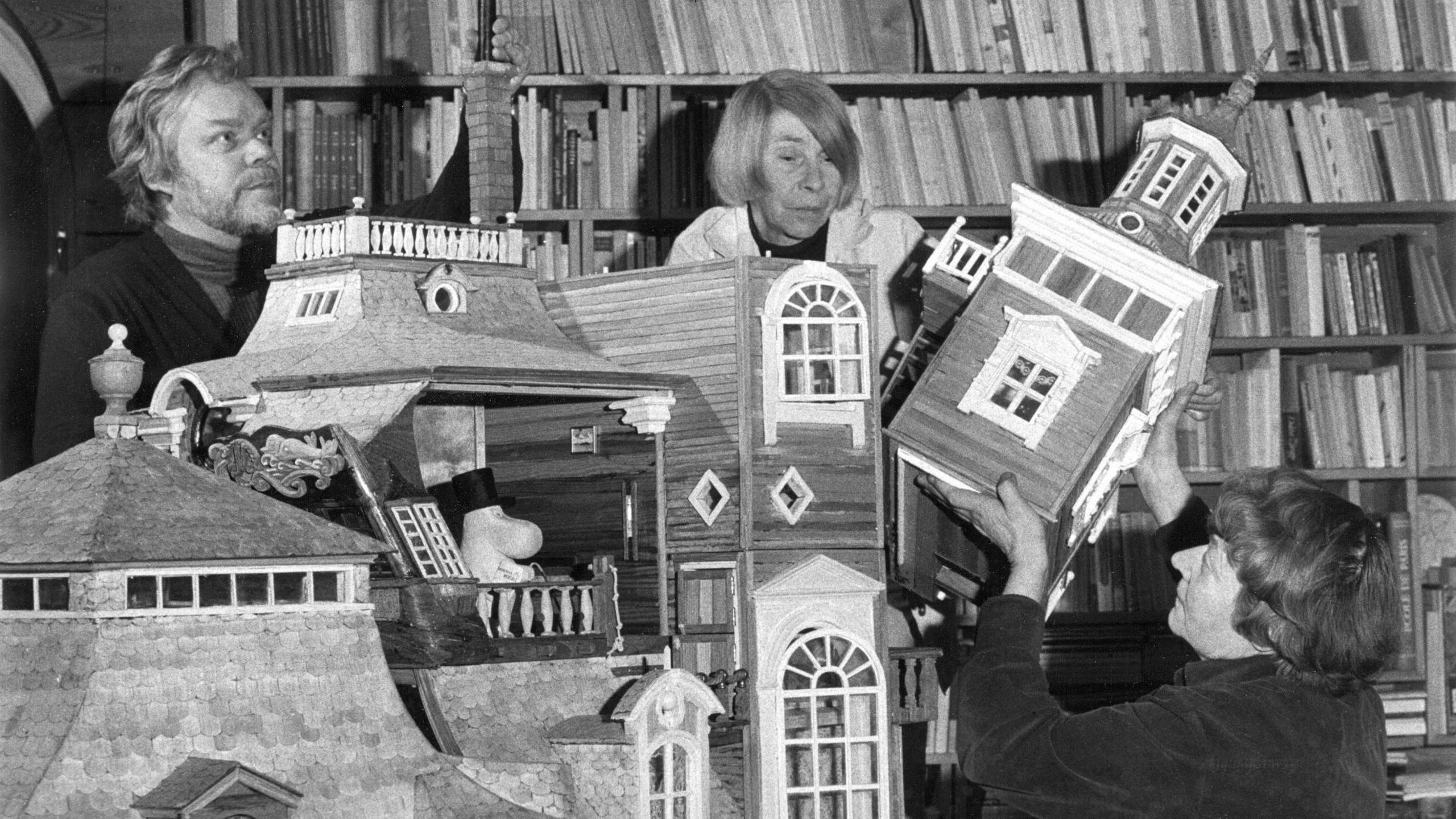 Black and white image of three people standing in front of bookshelves, including Tove Jansson, as they construct a large dolls' house sized Moomin house