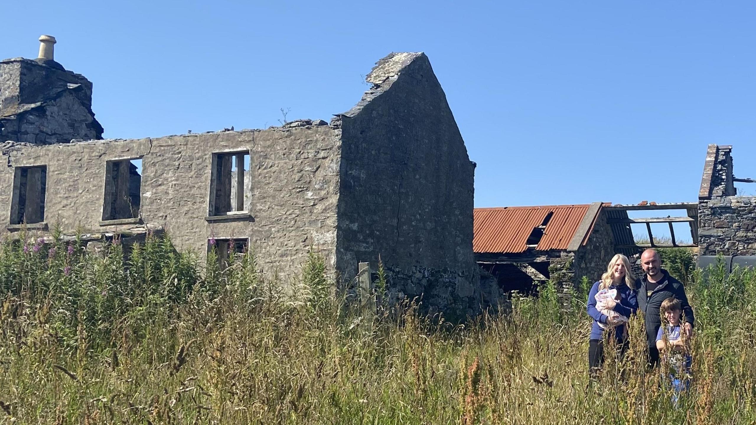 The family stood in overgrown weeds with a large roofless building and a barn behind with metal corregated roof, which is partially broken off.