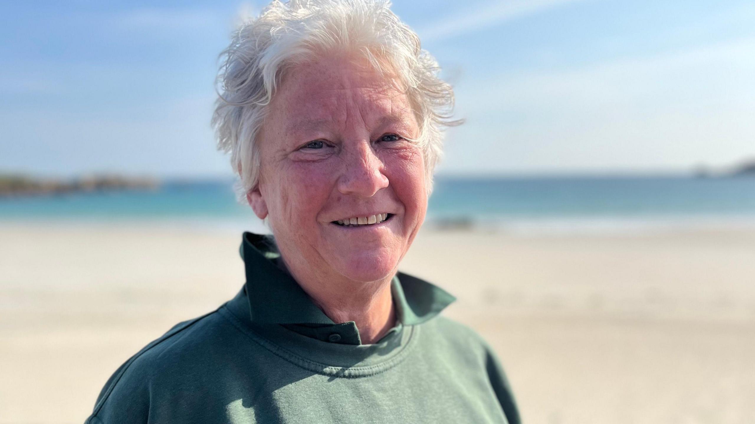 A lady in a green jumper with a green t-shirt with grey hair siling towards the camera.