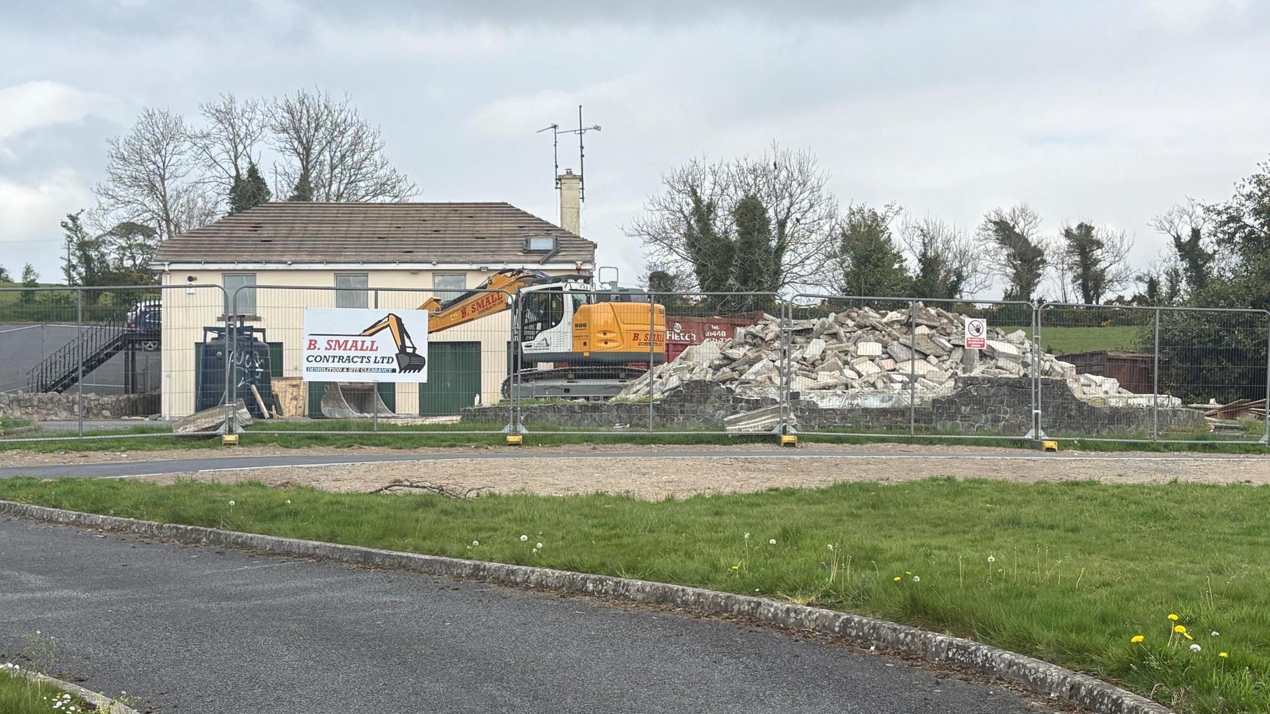 The former site of Clonduff parochial house. A large pile of rubble sits where the main building used to stand. A metal safety fence is blocking off the whole site. Roof tiles have been stripped from a smaller parish building in the background.