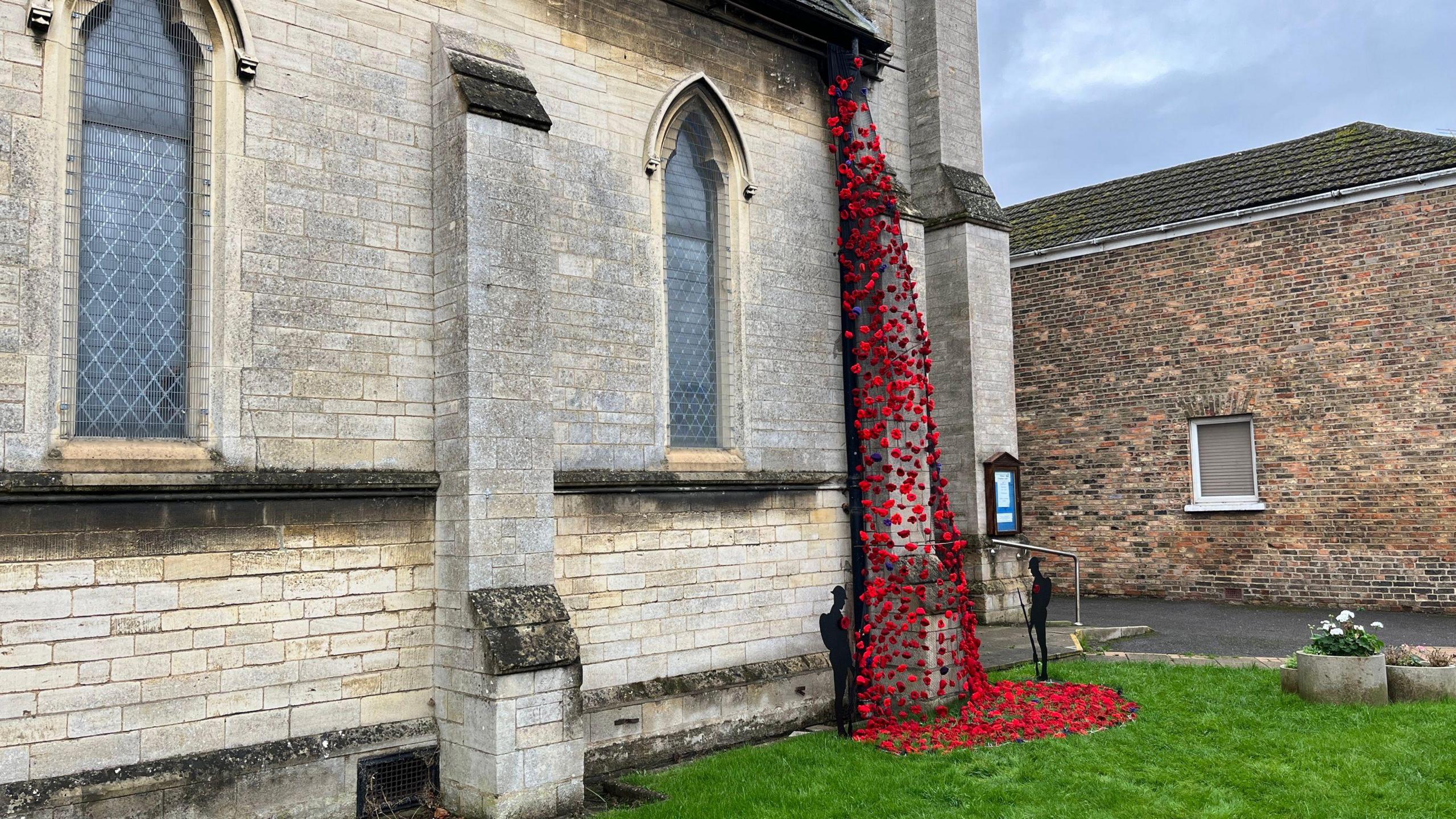 One side of St Matthew's Church in Eye can be seen. It has light, sandstone-coloured bricks, arched windows and buttresses, one of which is covered with a black net onto which is attached red and purple knitted poppies. These are also pooled on the grass at the bottom of the cascade alongside two black metal soldier figures. To the right of the image is part of another, red-brick, building. 