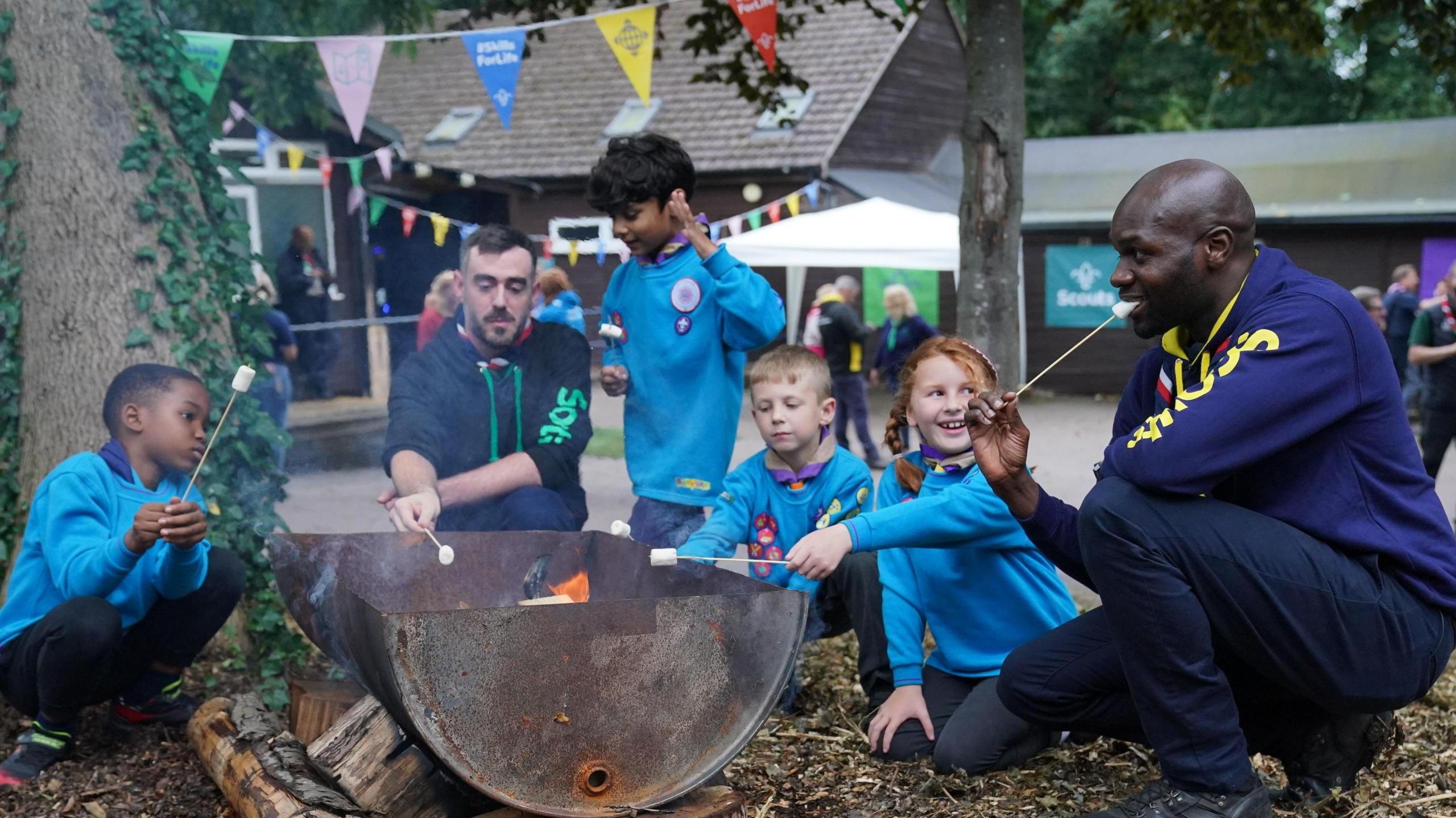 Chief scout Dwayne Fields toasts marshmallows over a fire with four young Scouts. He is kneeling down to the right of the fire with the children sat beside him.