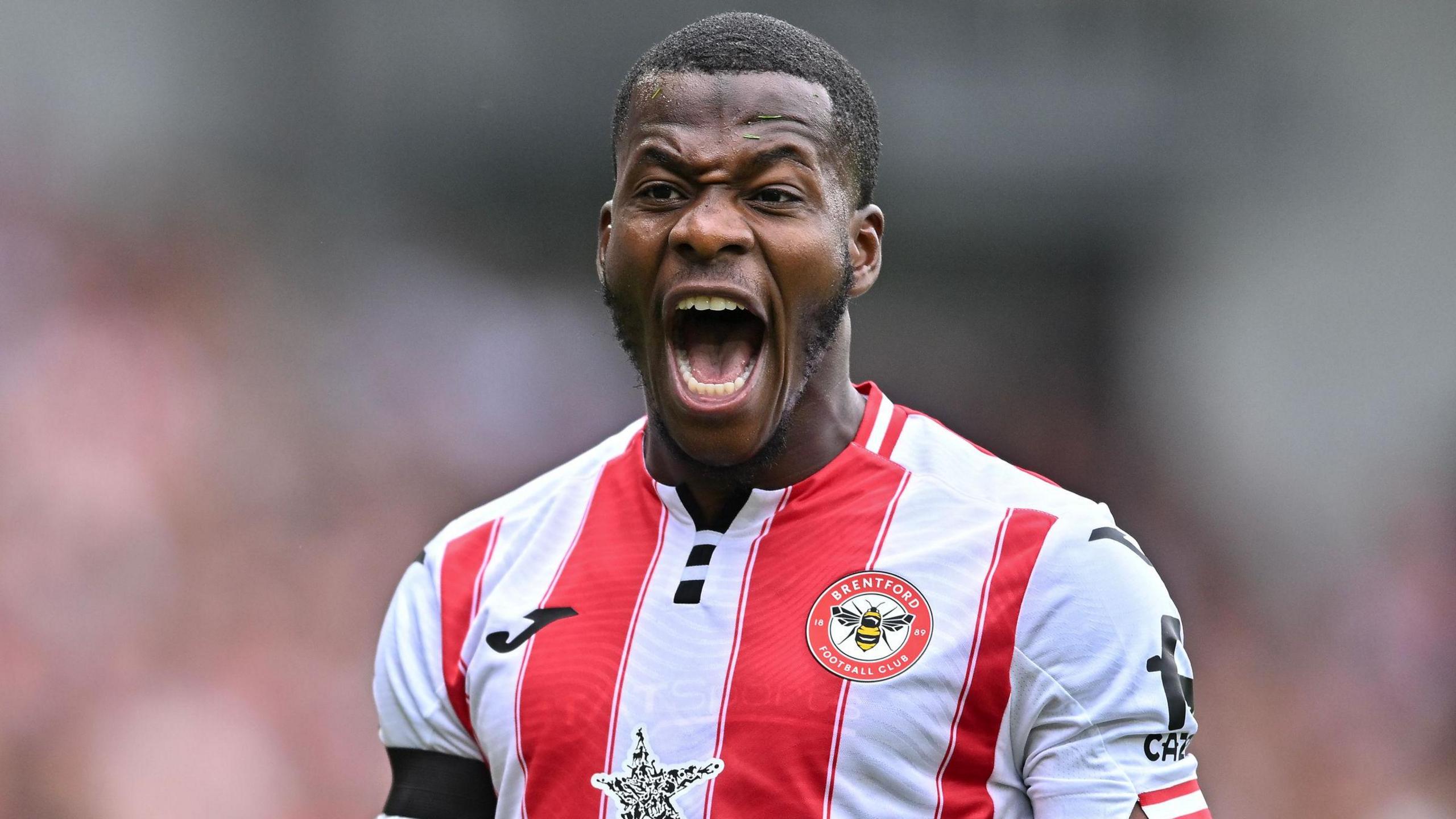 Dango Ouattara shown from chest up playing for Brentford Football Club in the home jersey with wide red and white vertical stripes. The forward is opening his mouth very wide to shout, with his teeth and tongue visible