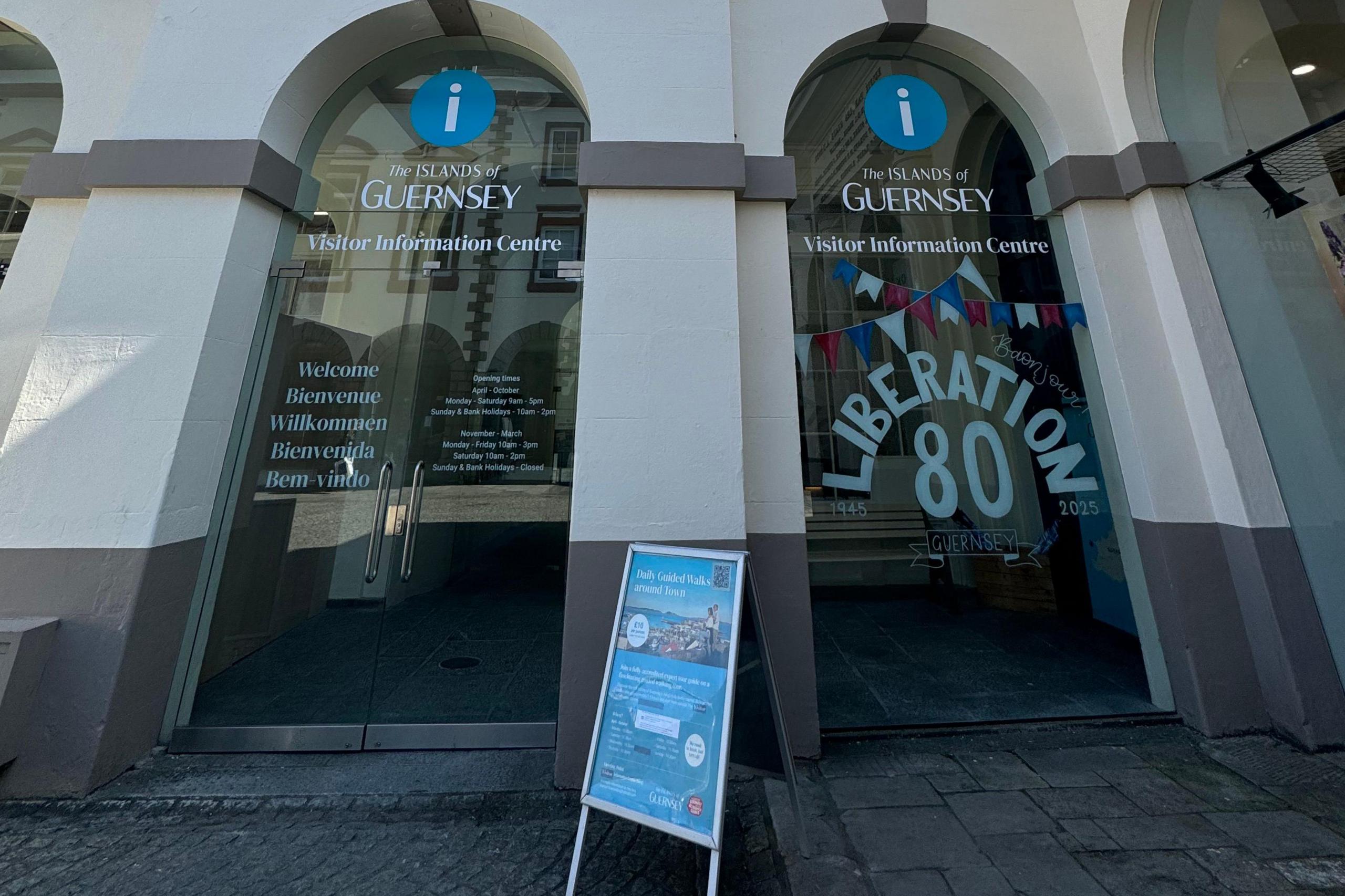 The photo shows the entrance to Guernsey Visitor Information Centre.  It features two large glass doors. The left door displays multilingual greetings and seasonal opening times. The right door shows the centre's name and a prominent “LIBERATION 80” graphic with the years “1945” and “2025” marking the 80th anniversary of Guernsey's Liberation from occupying rule on May 9th 1945. An A-board sign stands outside promoting daily guided walks around Guernsey's capital, St Peter Port. 