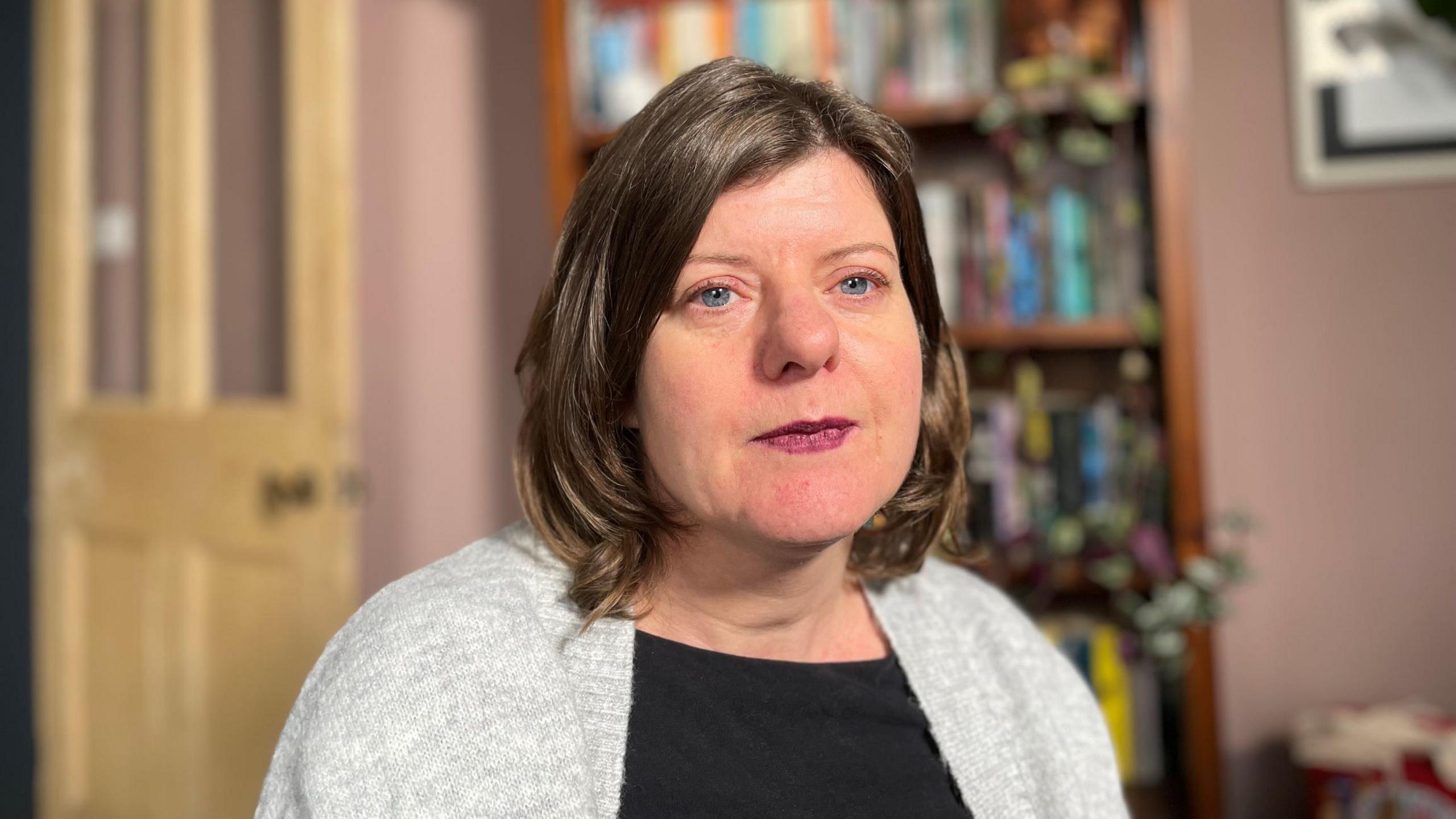 Sandy Brindley, with a black top and grey cardigan on. She is looking straight in the camera with a bookcase behind her