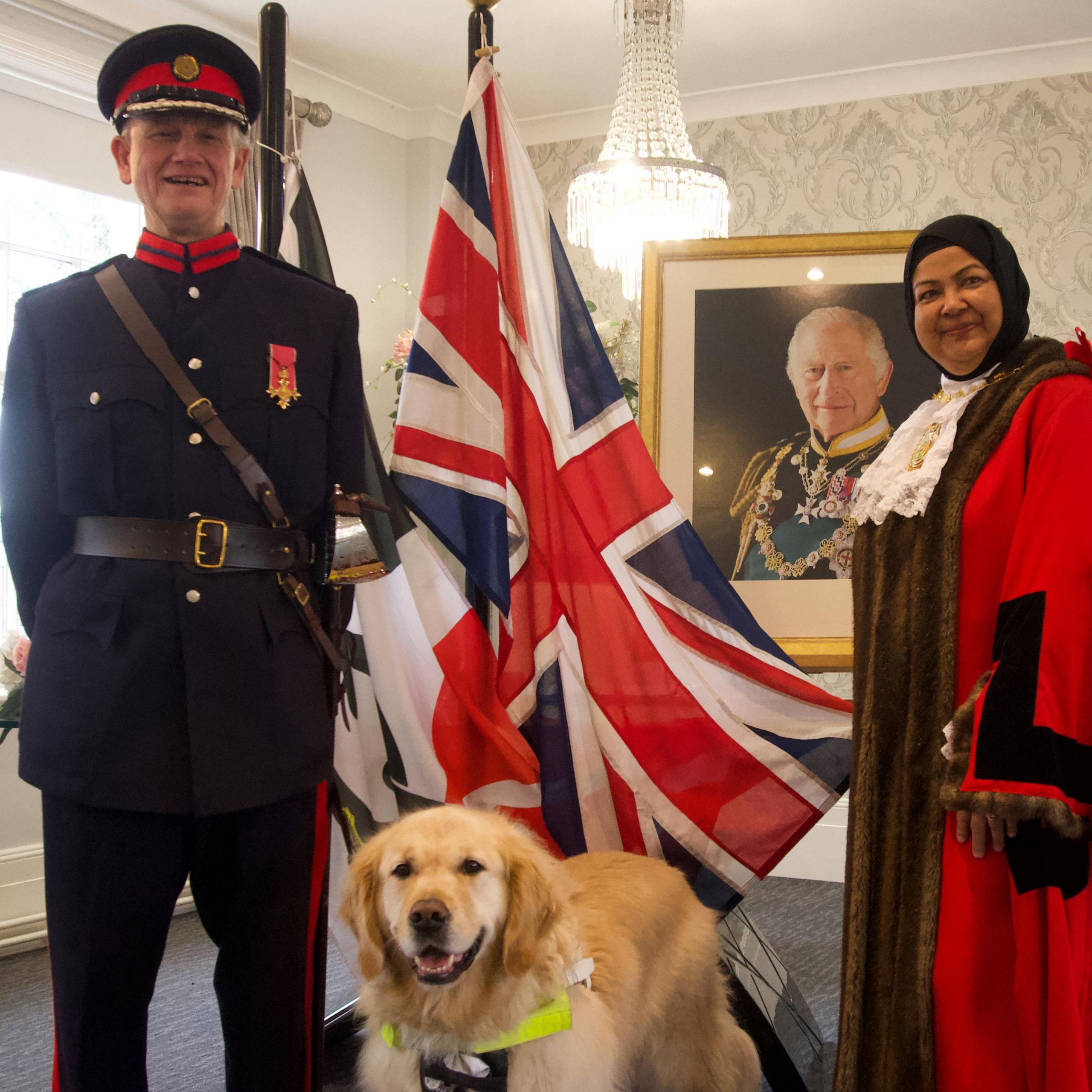 A man in navy blue military uniform with a brown leather belt and a navy and red peaked cap. He is wearing an OBE medal and is standing next to the Black Country flag and Union flag with his golden retriever guide dog in the foreground and a female mayor in her red and brown robes to his left. In the background is a photograph of King Charles. 