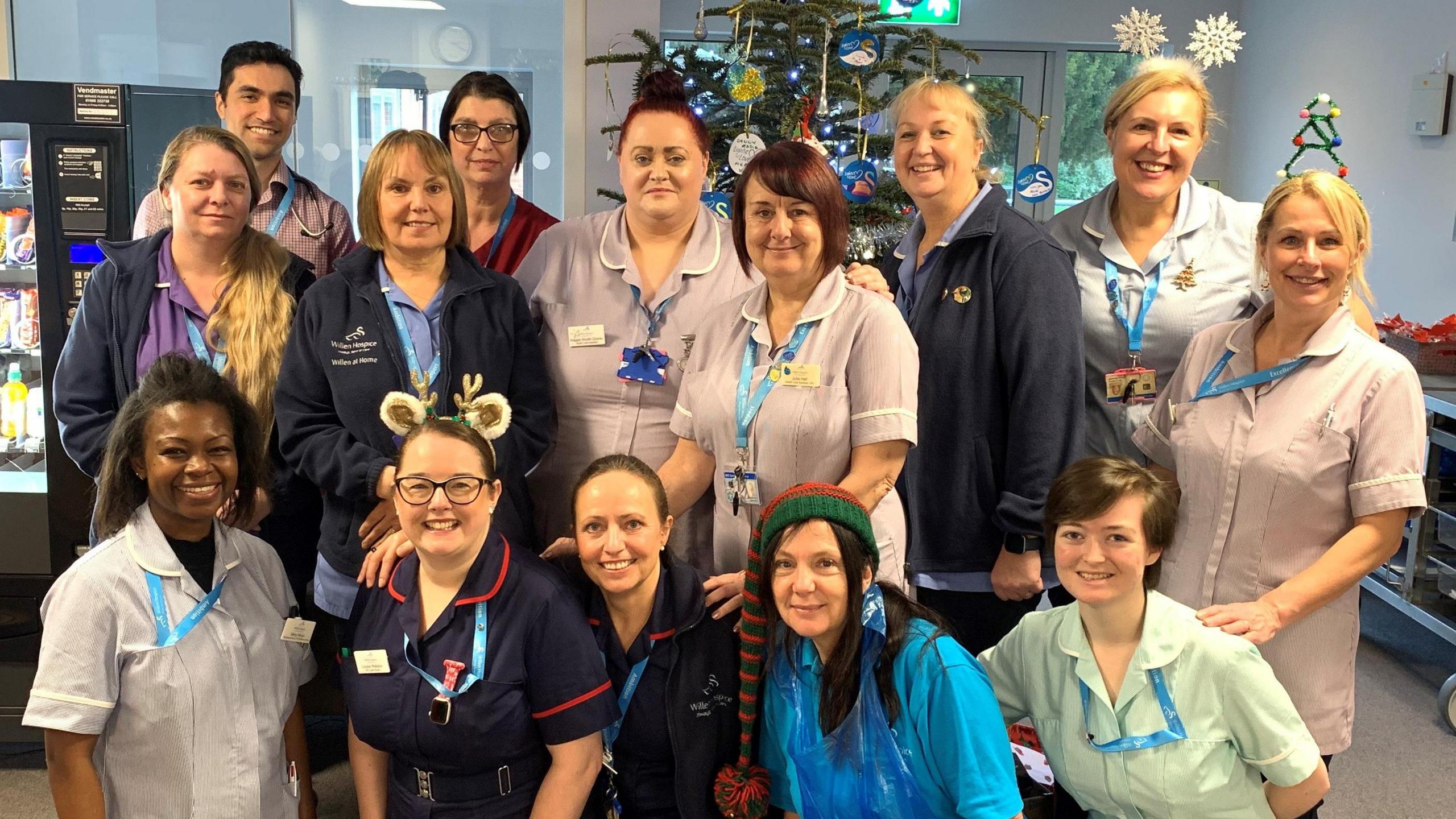 A team photo of staff at the inpatient unit at Willen Hospice. There are 14 people in the photo, wearing nursing uniform and blue lanyards. Some wear elf hats and headbands, and they are all smiling. There is one man and the rest are women. A Christmas tree is in the background and it looks like they are in the lobby with a vending machine to the right. 