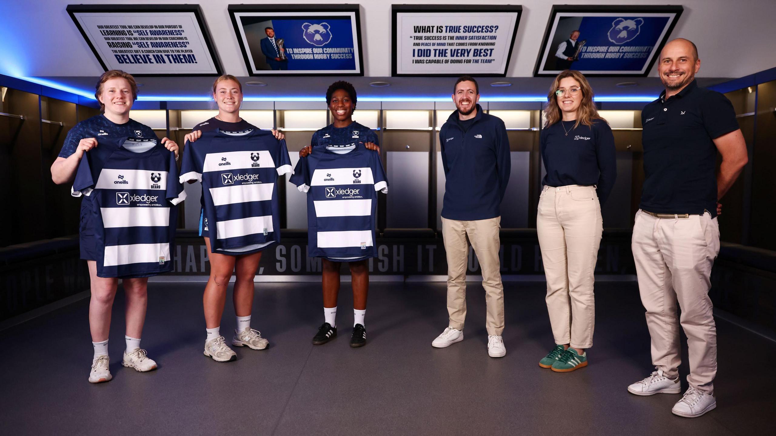 A group of Bristol Bears women players hold up the club's home shirt with the emblem of sponsor Xledger on it. Staff from Xledger are also in the picture, which is taken in the dressing room at Ashton Gate.