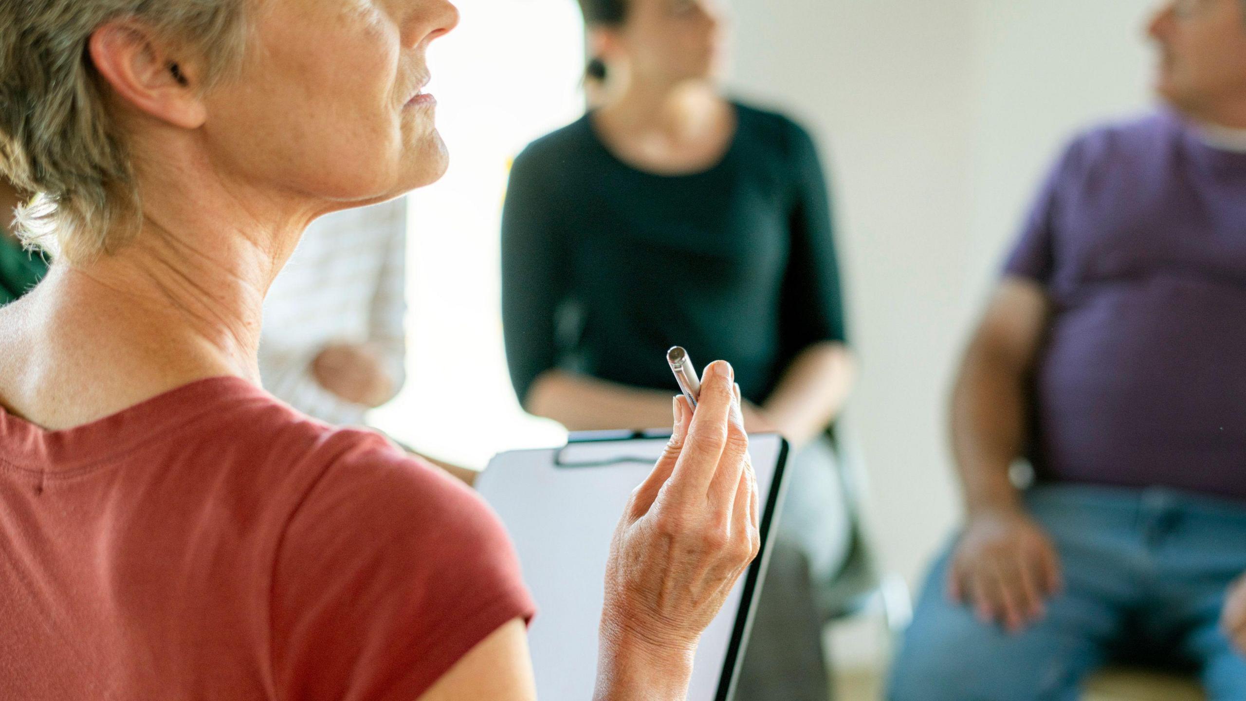 A woman holds a clipboard in front of people sitting on chairs. 