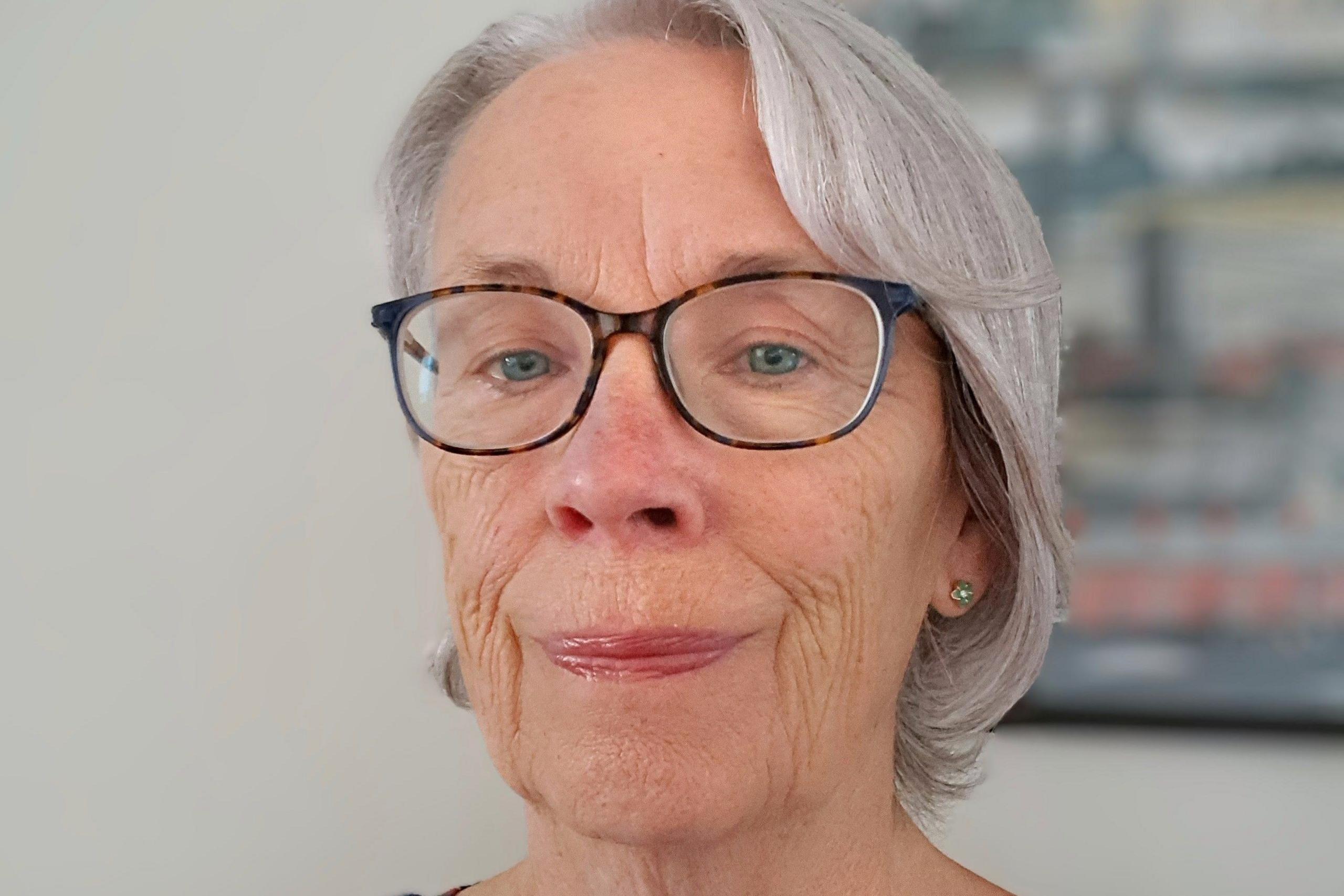 Jacqueline Gantley is wearing dark rimmed glasses and has chin-length grey hair. She is standing against a white wall with a picture on the wall.