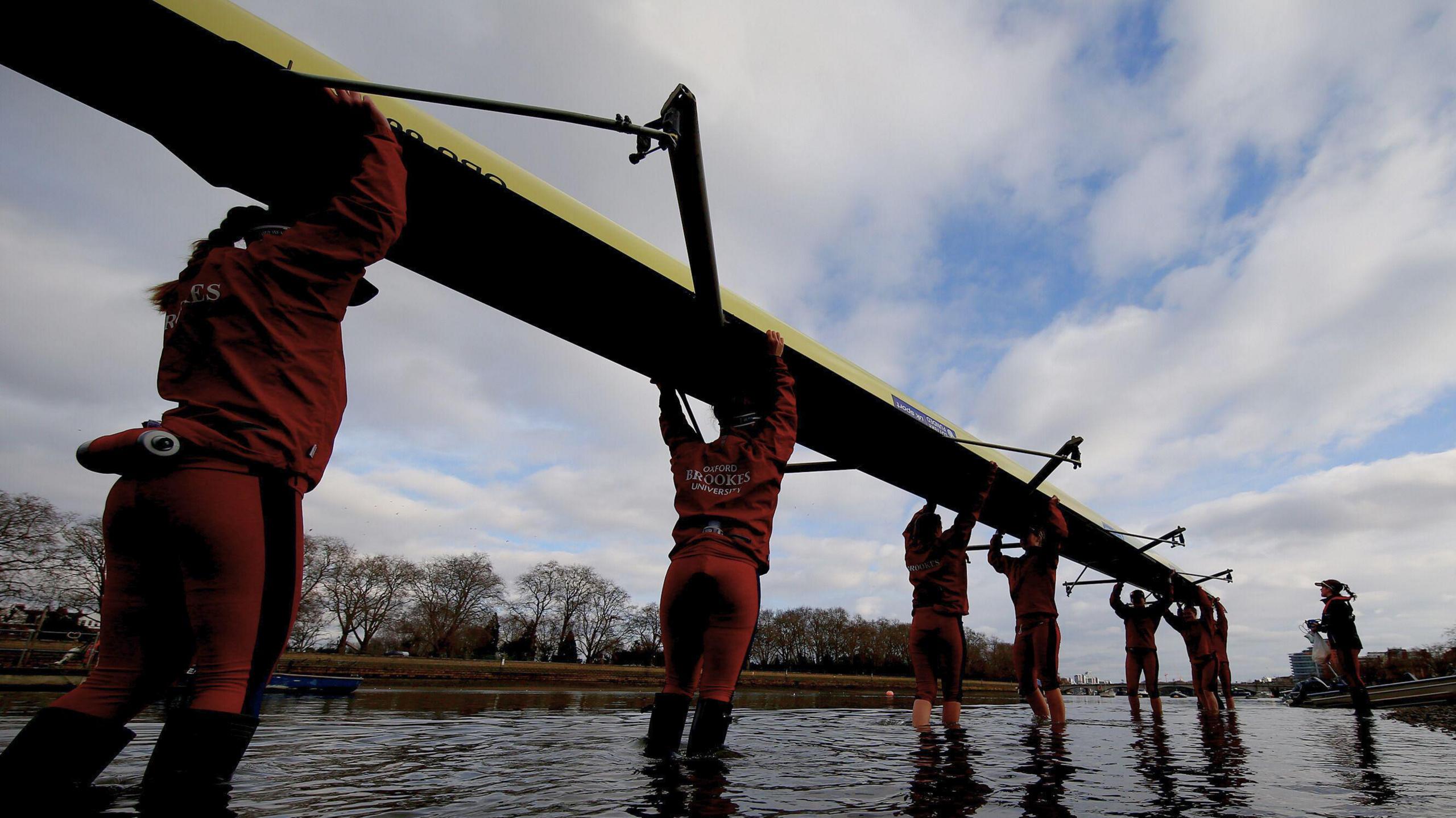 Seven women are holding a rowing boat above their heads. They're wading into a river as they carry the boat. They're wearing matching sports kits, which show they're rowers for Oxford Brookes rowing team. The sports jackets and trousers are red. The sky is blue and cloudy.