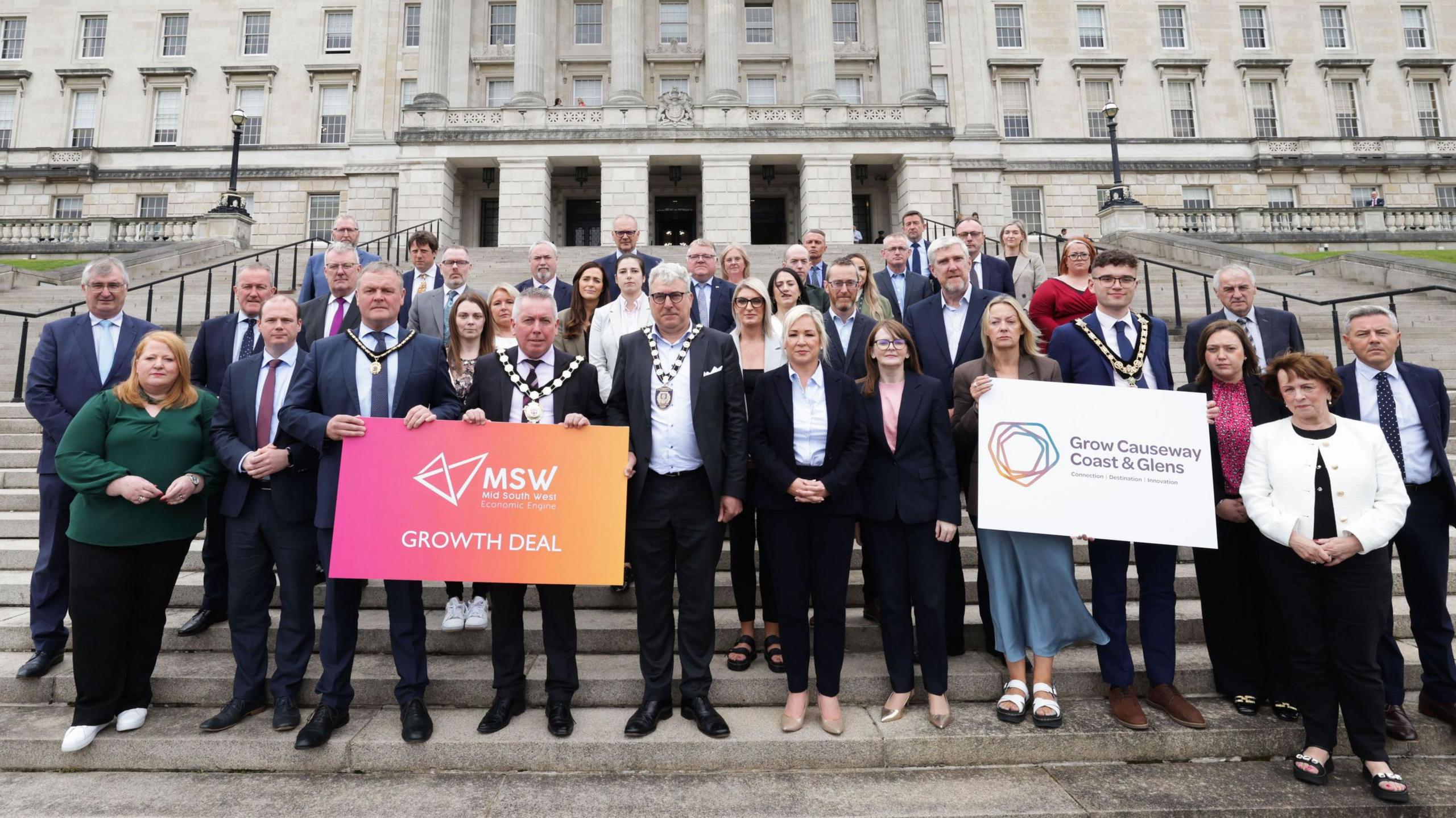 A group of people wearing suits standing on the steps in front of Stormont, which is a white building with 6 pillars on the front and lots of square windows.