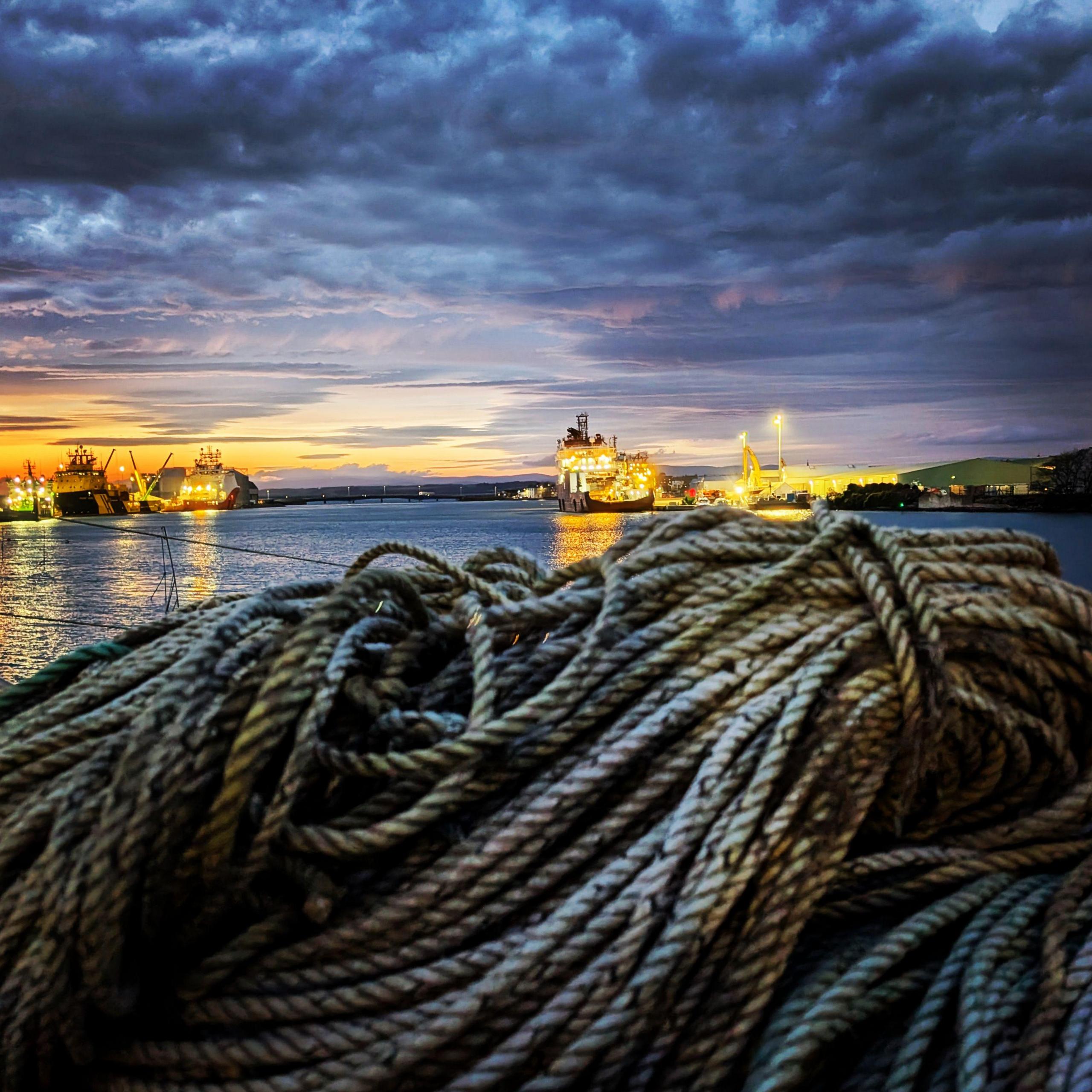 Fishing ropes in the foreground of a photo taken at a port. Ships with bright lights on are in the background.