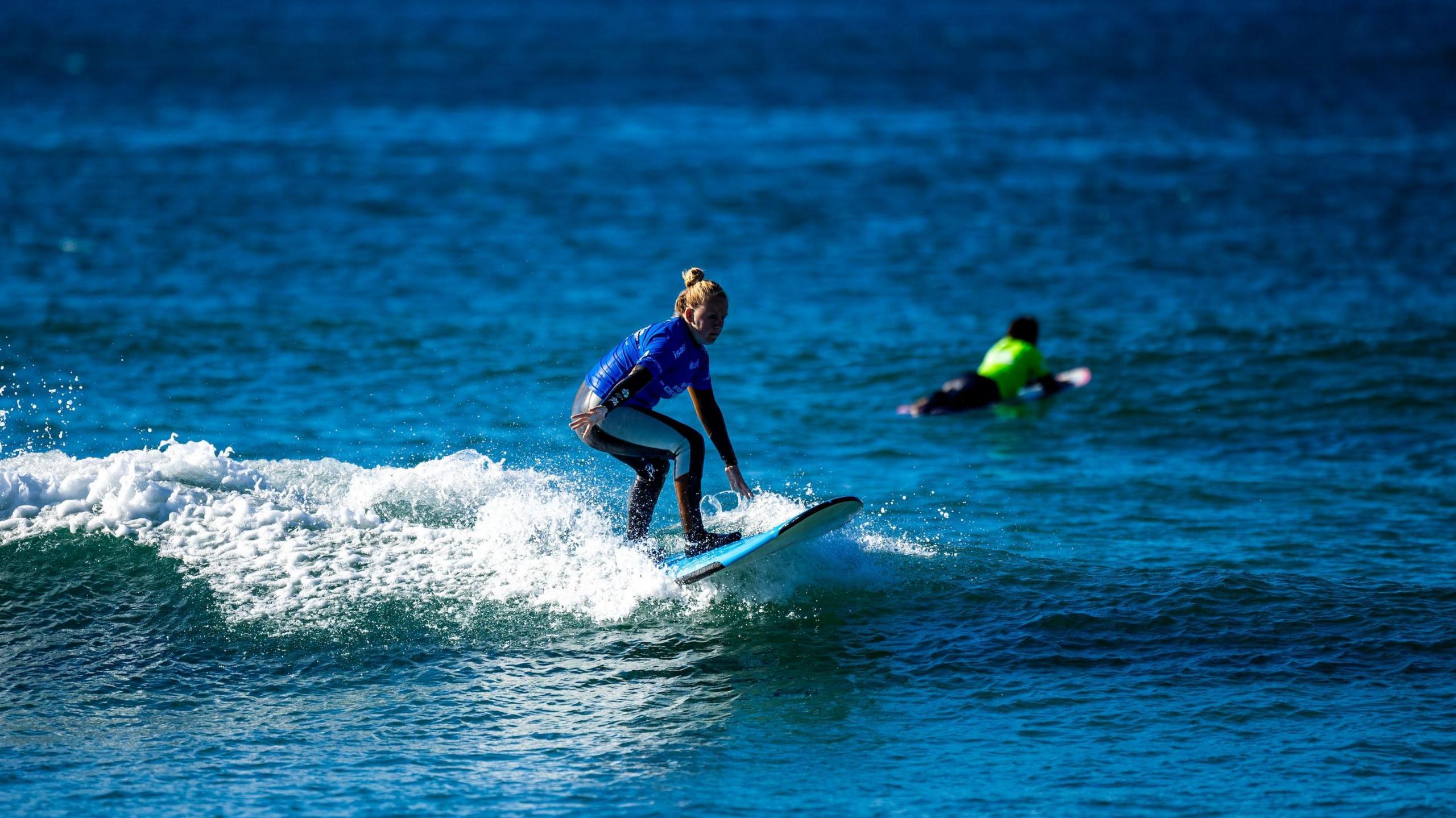 A lady is surfing on water. She has blonde hair and is wearing a blue and black wetsuit. Her board is light blue and the water is of similar colour.