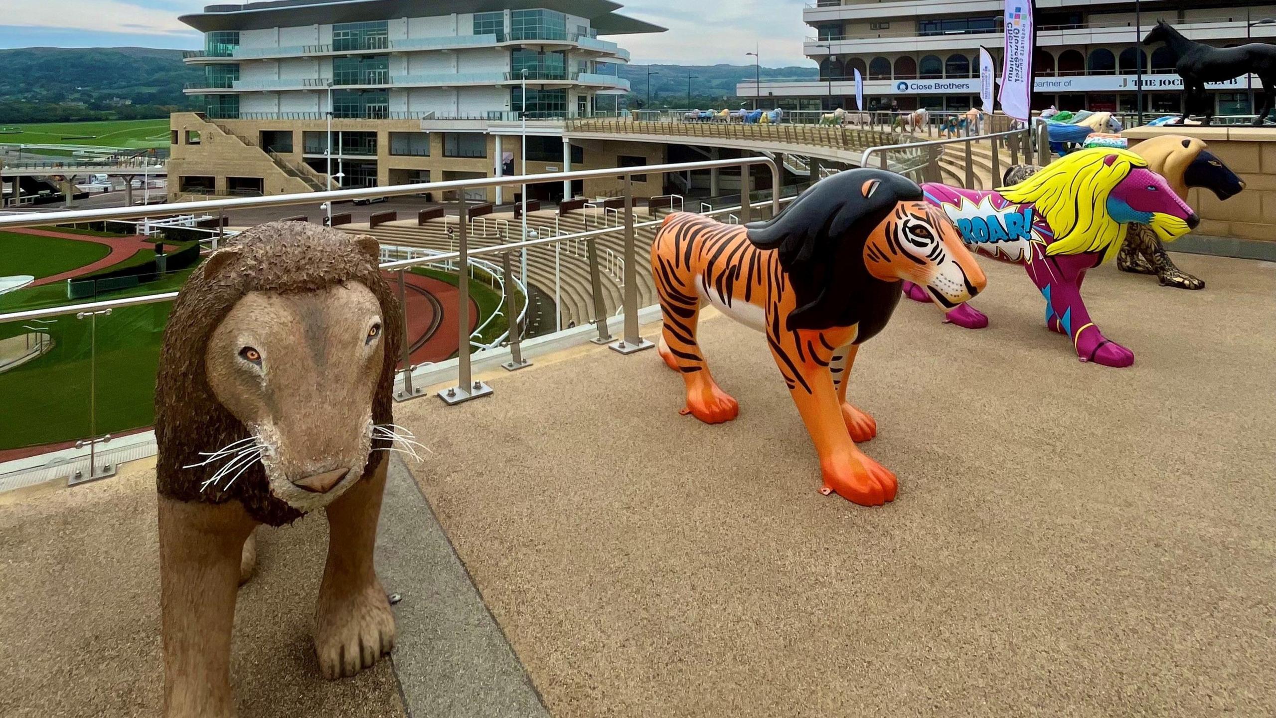 Four lion statues lined up overlooking Cheltenham Racecourse. The nearest is brown and getting further away, they are painted orange (with black main), purple (with yellow main) and black (with gold main).