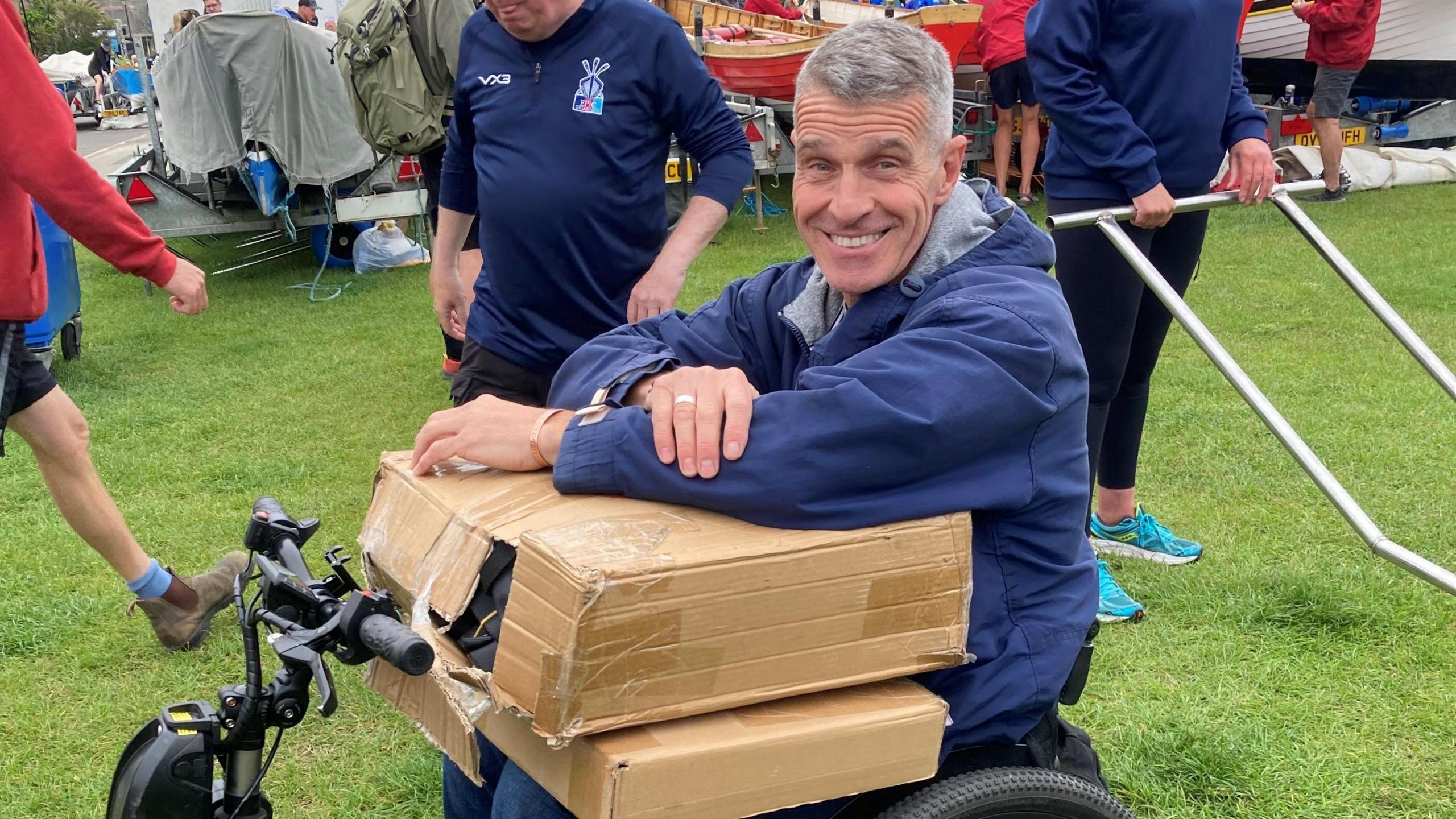 A man in a wheelchair smiles at the camera holding a box of kit for gig rowing.