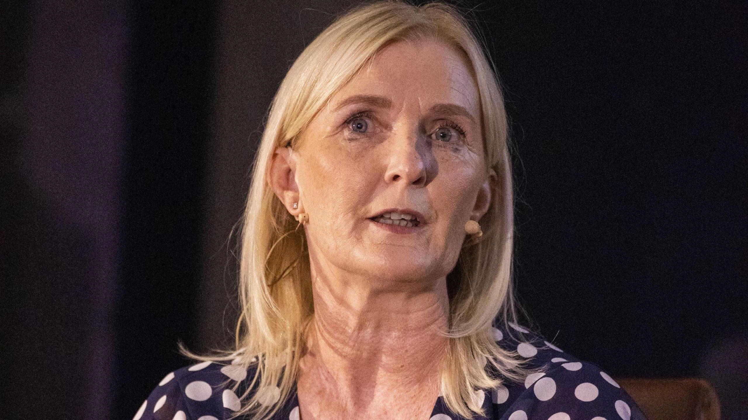 A long-blonde haired woman with a microphone on her left cheek, wears a navy dress with white polka dots. She has a small diamond earing in her left ear.