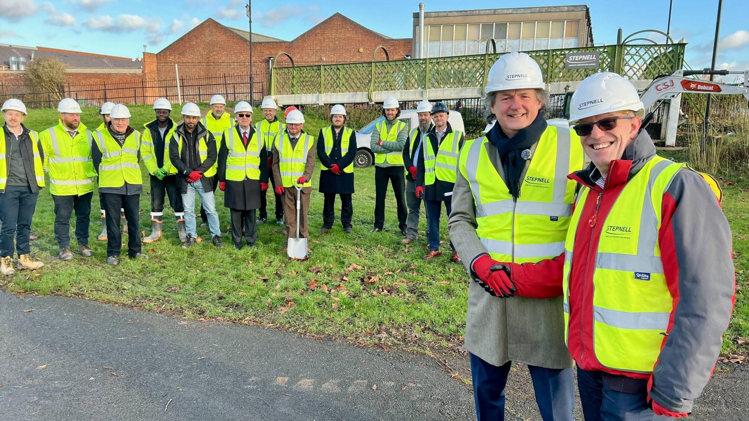 Fifteen men, all dressed in high-vis vests, red gloves and white hard hats stand on the grass beside an old steel bridge painted green. One of those men in the centre holds a spade. Two men dressed in the same attire are stood in front of them, shaking hands.  On the other side of the bridge are industrial buildings made of brick.
