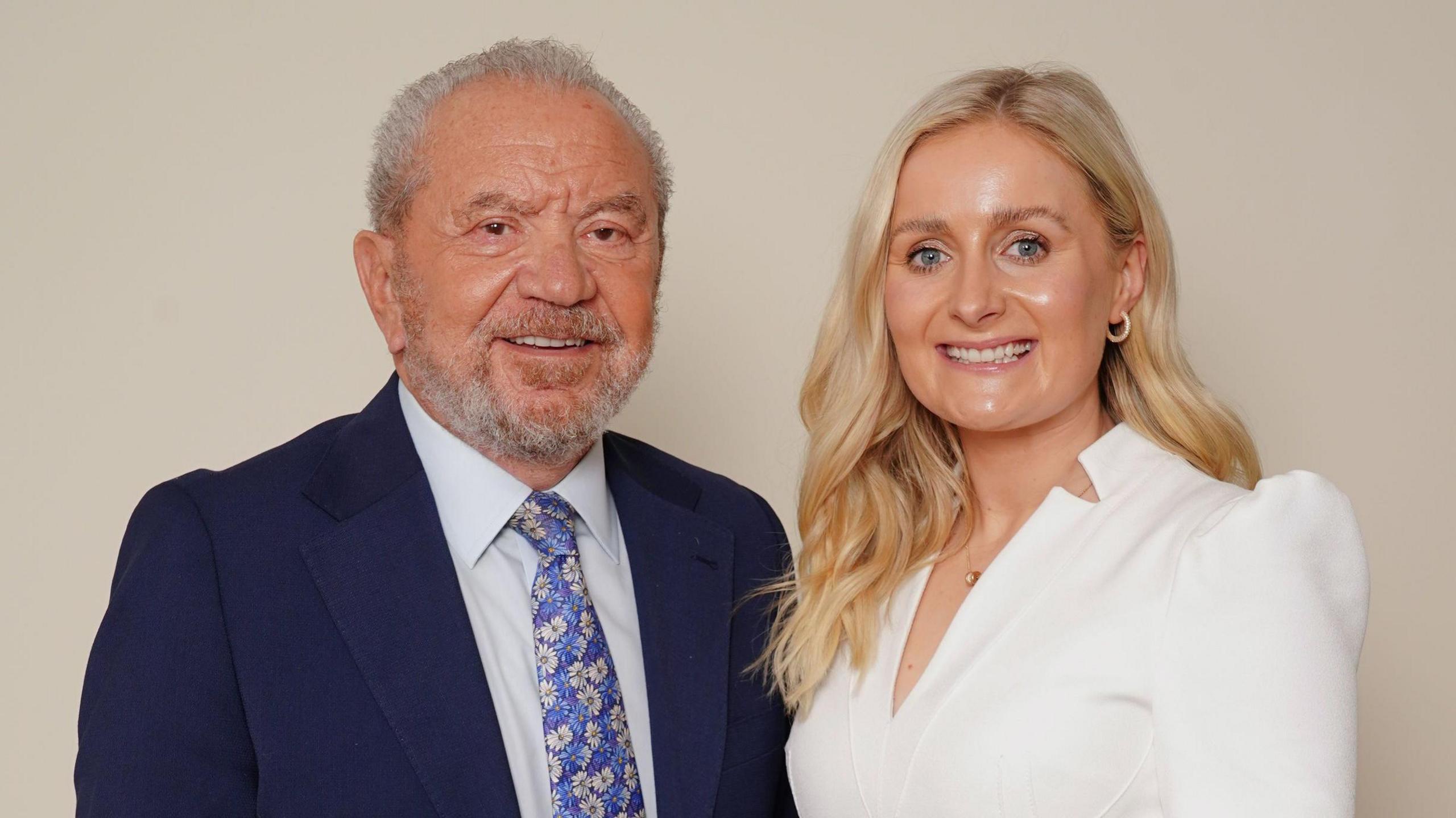 Lord Sugar, dressed in a suit, smiles next to Rachel Woolford, in a white suit.