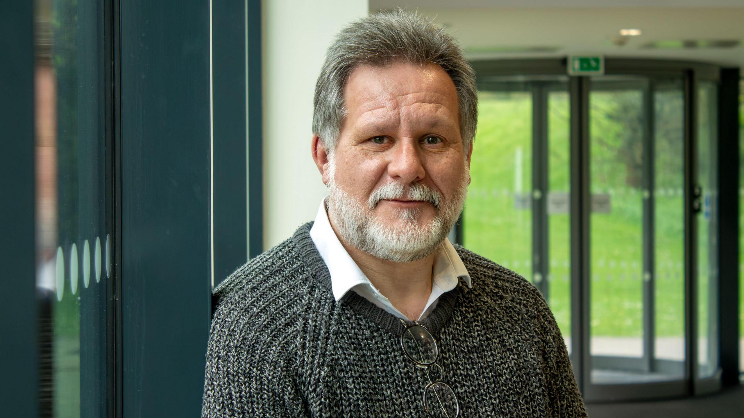 Matthew Wills wearing a white shirt underneath a knitted grey jumper, with his glasses dangling from the collar. He has thick, dark grey hair and a well trimmed light grey beard. He is sitting in the entrance foyer of a university, with a glass revolving door in the background that leads to a bright green grassy verge.