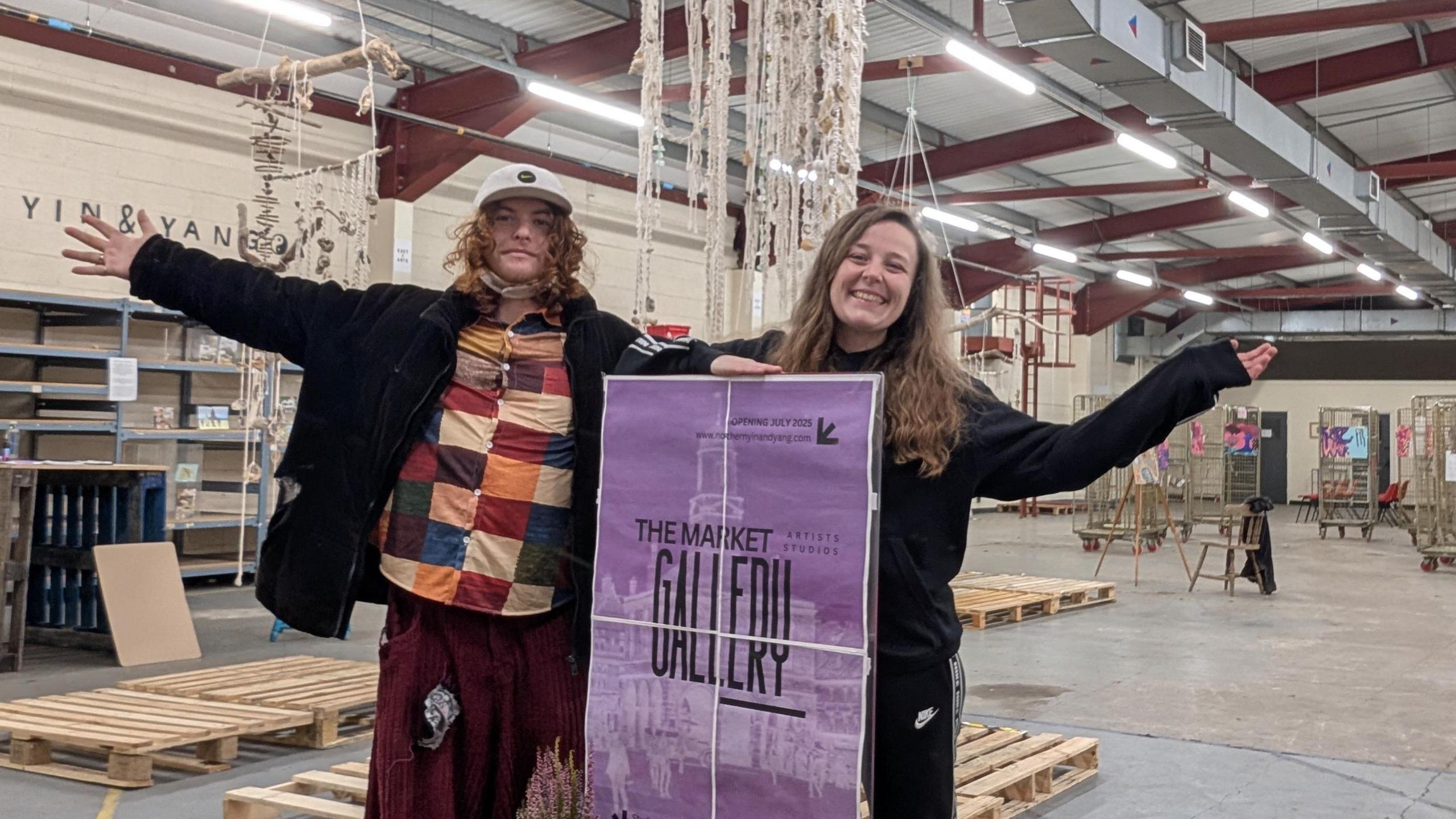 Theo Godfrey-Cass and Laura Robertson inside their new exhibit in the storage area of the former Wilko store. They are holding a purple sign with the name of the gallery on. Behind them are a number of pieces of art work on display.