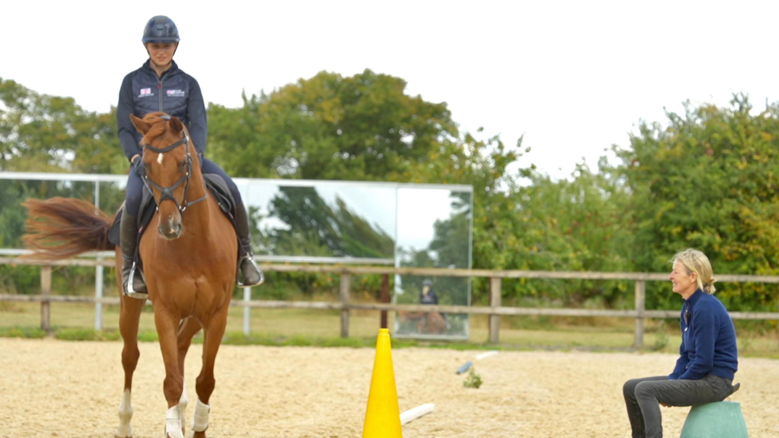 Ella rides Spice in the outdoor arena. She rides past her mother who is sitting on an upside down blue bucket in the school. A yellow cone rests in between them as well as a pole.