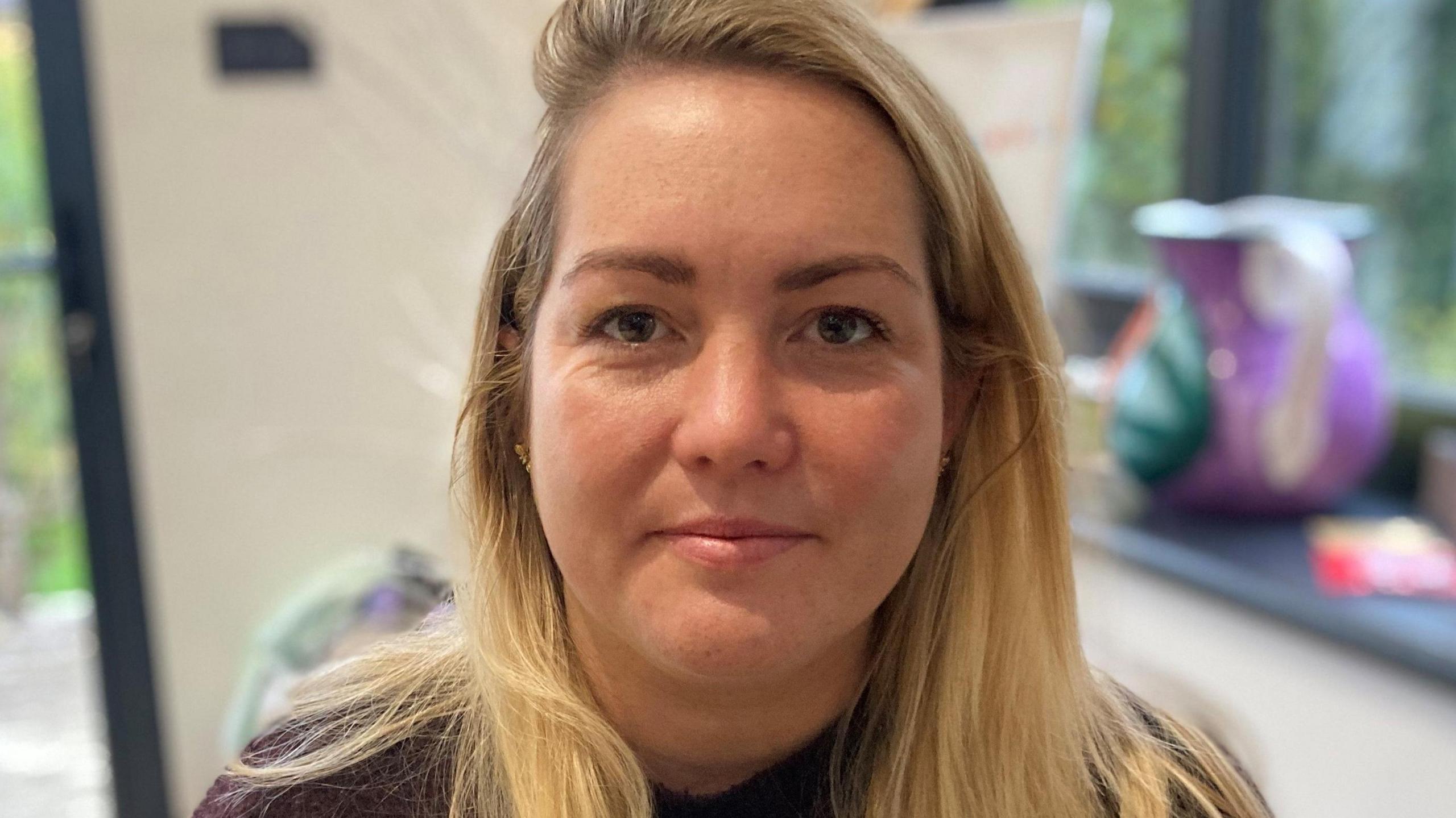 A woman with long blonde hair and hazel eyes looks directly at the camera. She is wearing a burgundy top. The background behind her is out of focus but shows a window with a jug in it and a glass door.