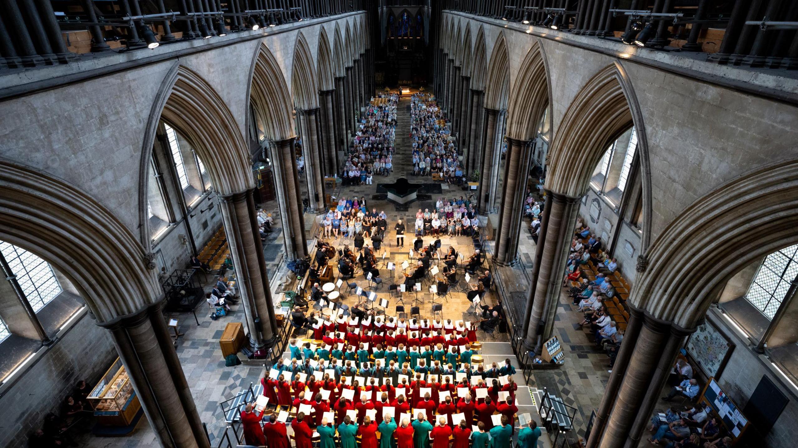 The interior of Salisbury Cathedral is seen in a picture taken from high up, with a choir in the foreground and the audience in the background. The cathedral's impressive structure dominates the image