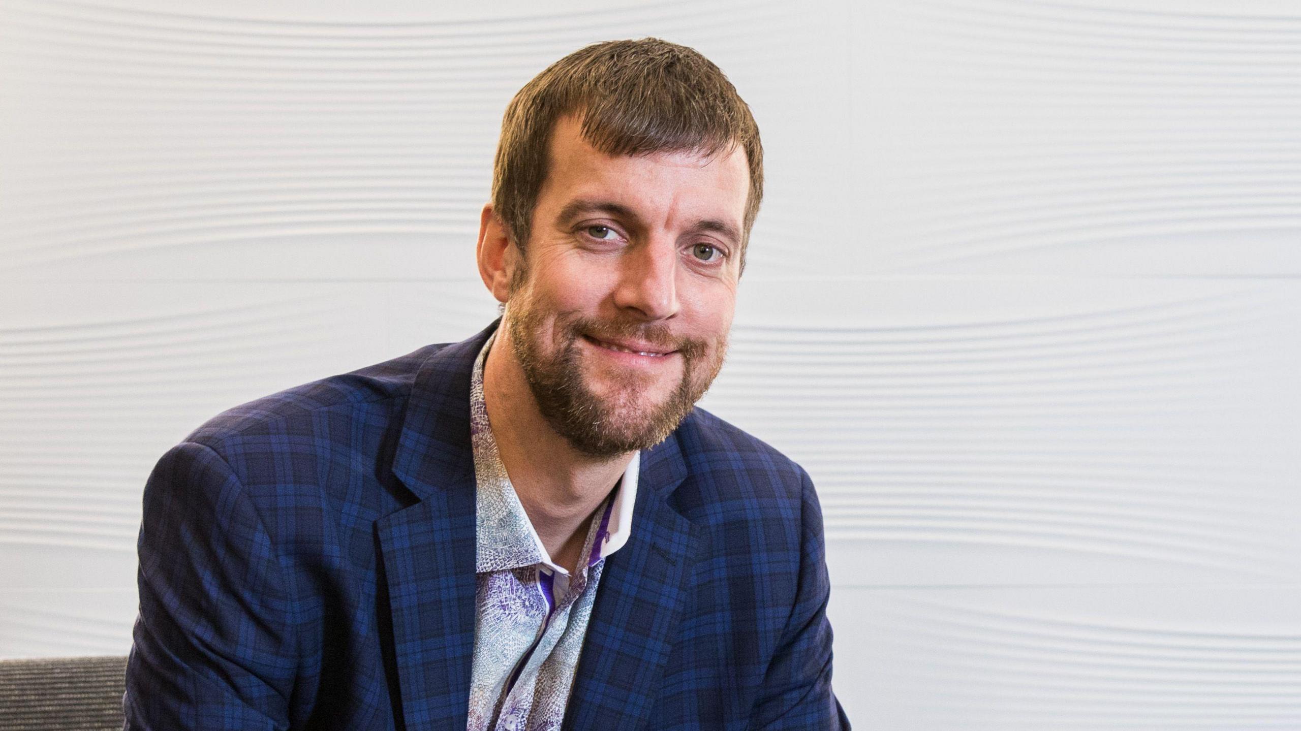 Headshot of Jason Trice smiling at the camera. He has brown hair and a brown beard and wears a shirt and jacket.