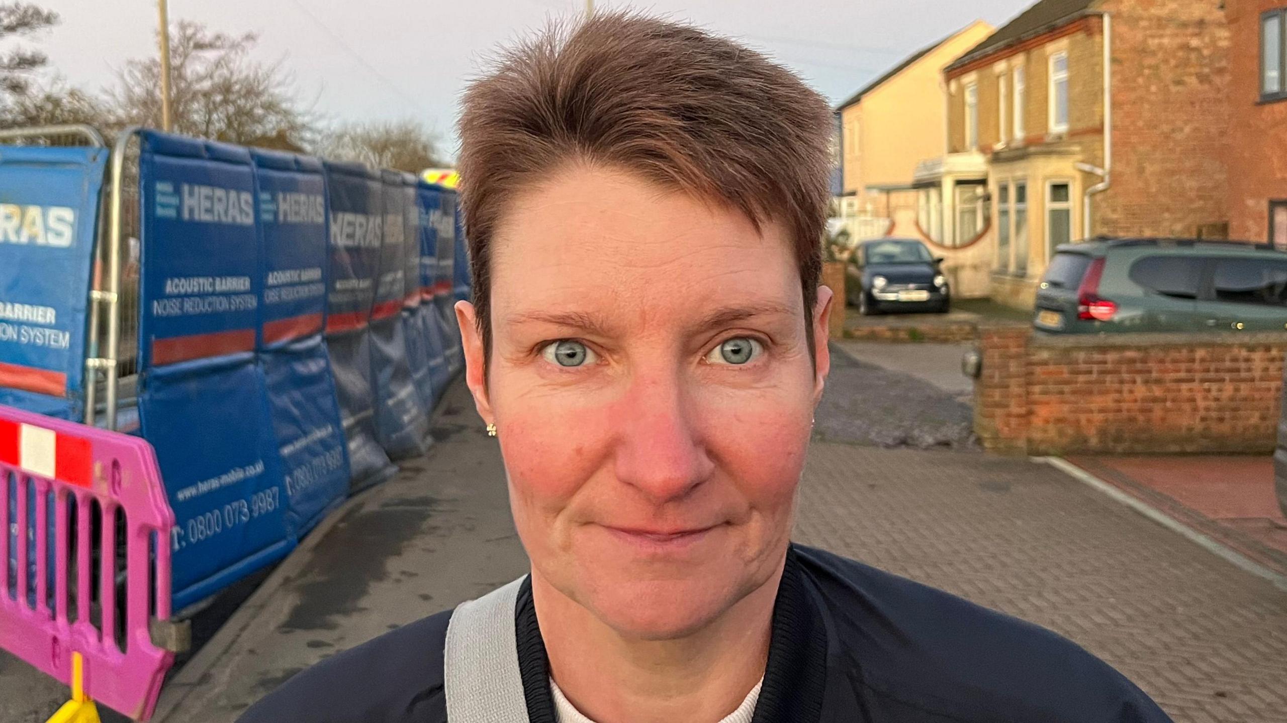 Zoe Ruddell looking straight into the camera while stood on the street. She has short dark hair and is wearing a black zipped jacket with a white T-shirt inside. There is a grey bag strap on her right shoulder. In the background is a cordoned off roadworks area and some cars parked on driveways.