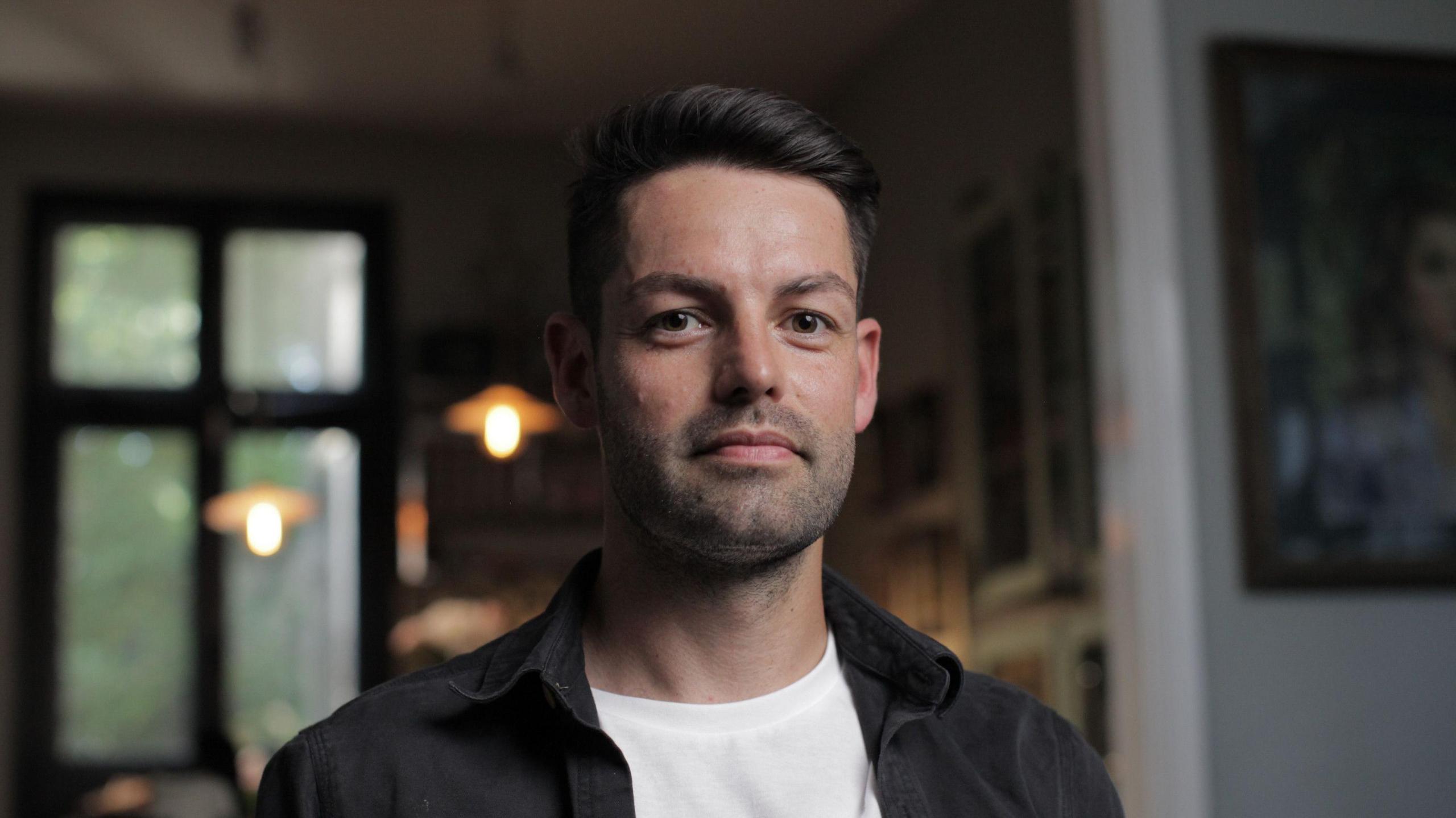 Louis Mullan pictured looking at the camera. He is wearing a dark grey overshirt with a white t-shirt. He has short, dark hair swept to one side and he has dark stubble. He is pictured in a house with paintings behind him.