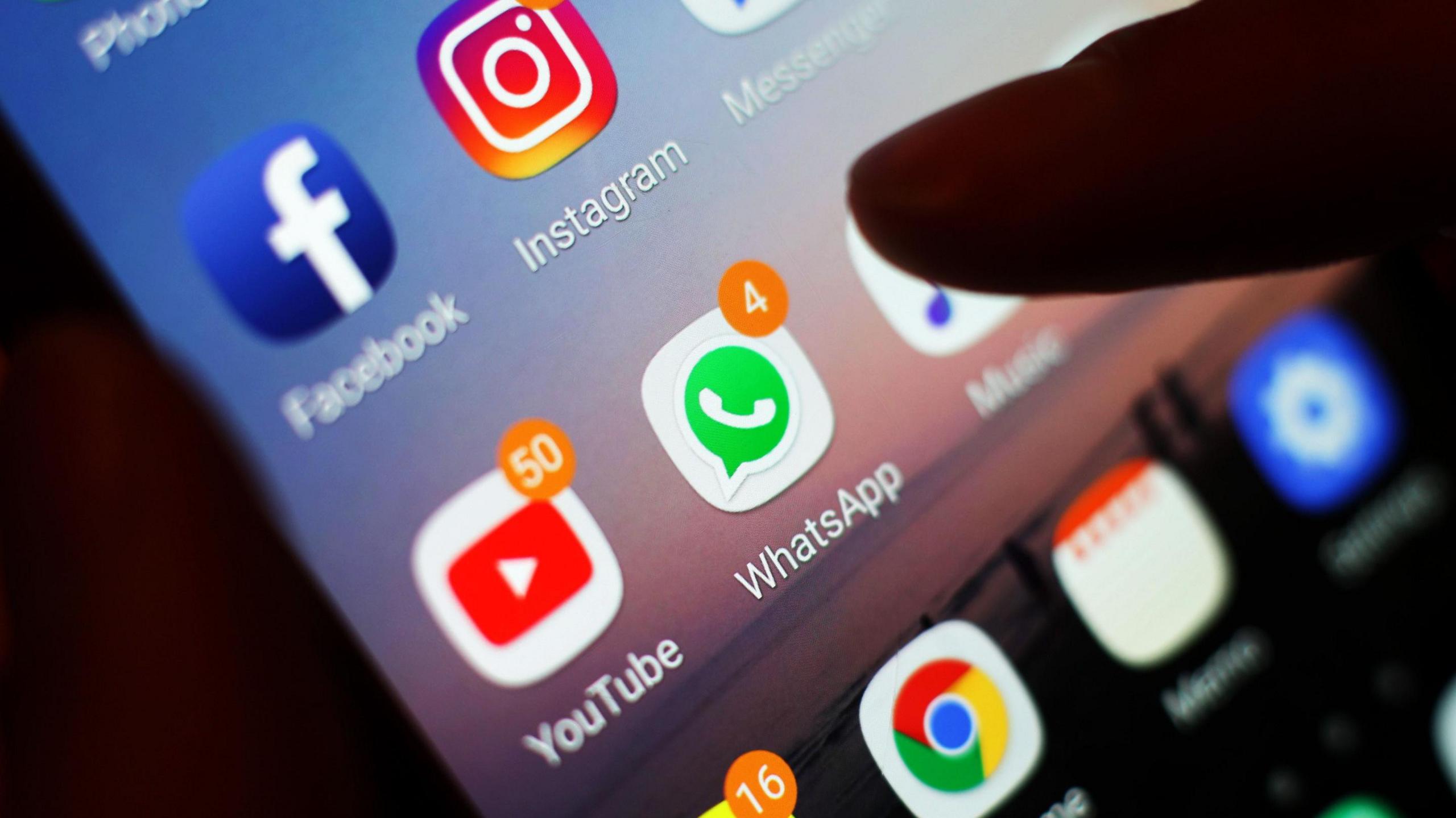 This is a close up photo of a screen. A finger is seen going to tap on a WhatsApp icon on a smart phone. There are several other apps on the screen, including YouTube, which has 50 notifications showing. 