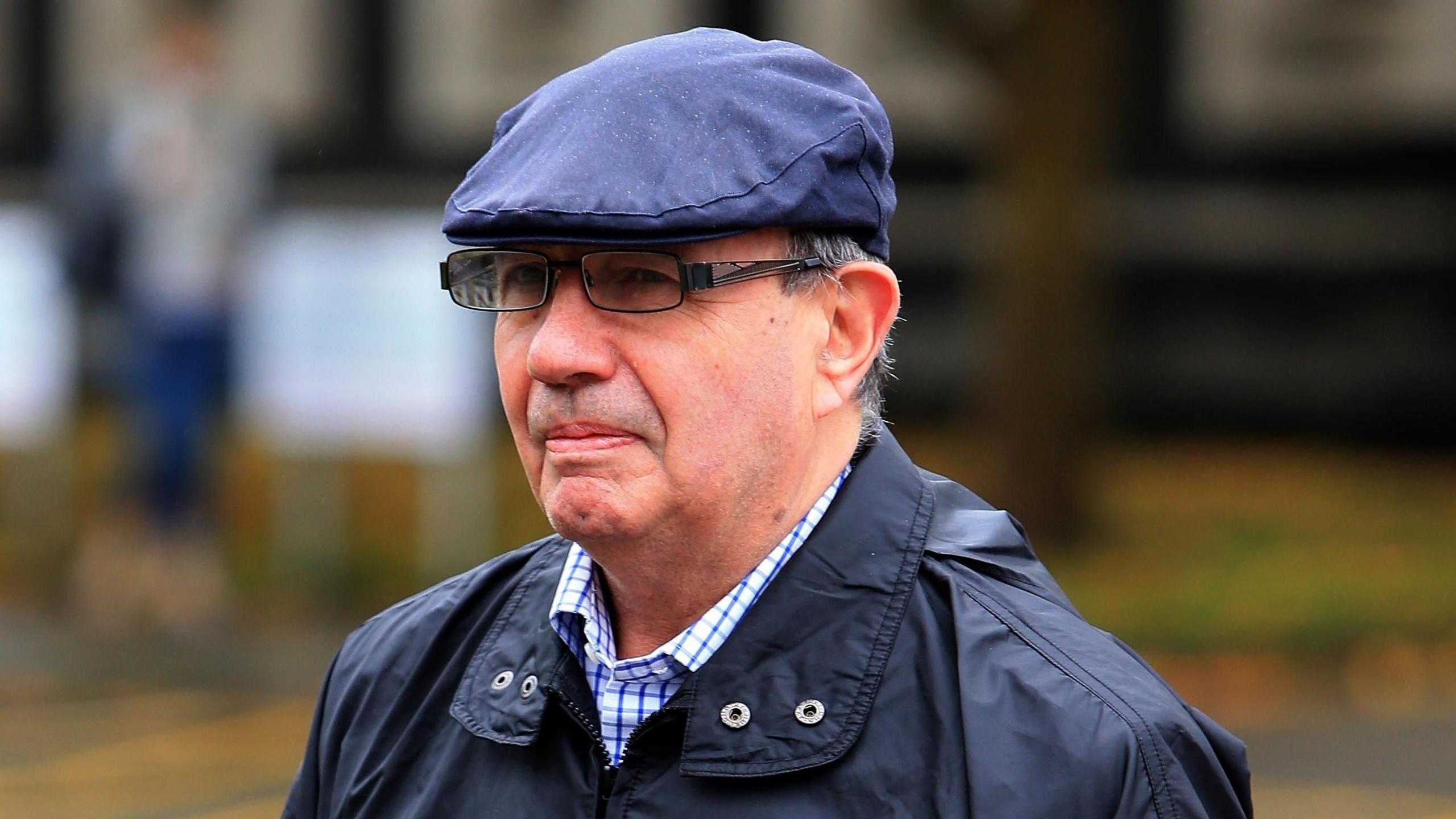 A man with black-rimmed glasses, wearing a blue flat cap, a blue jacket and a blue and white checked shirt underneath. He is walking into court, but only his shoulders and face can be seen in this image.