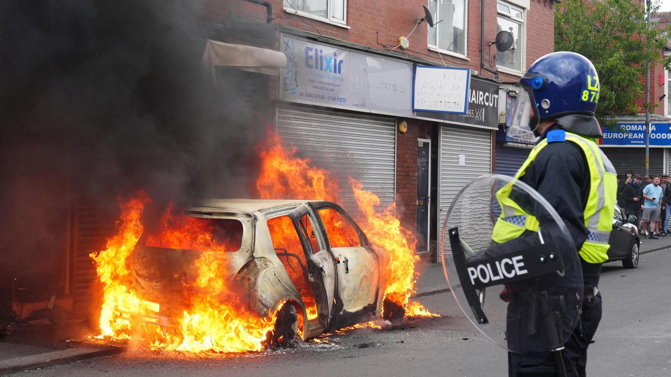 A police officer in riot gear looks at a burning car.