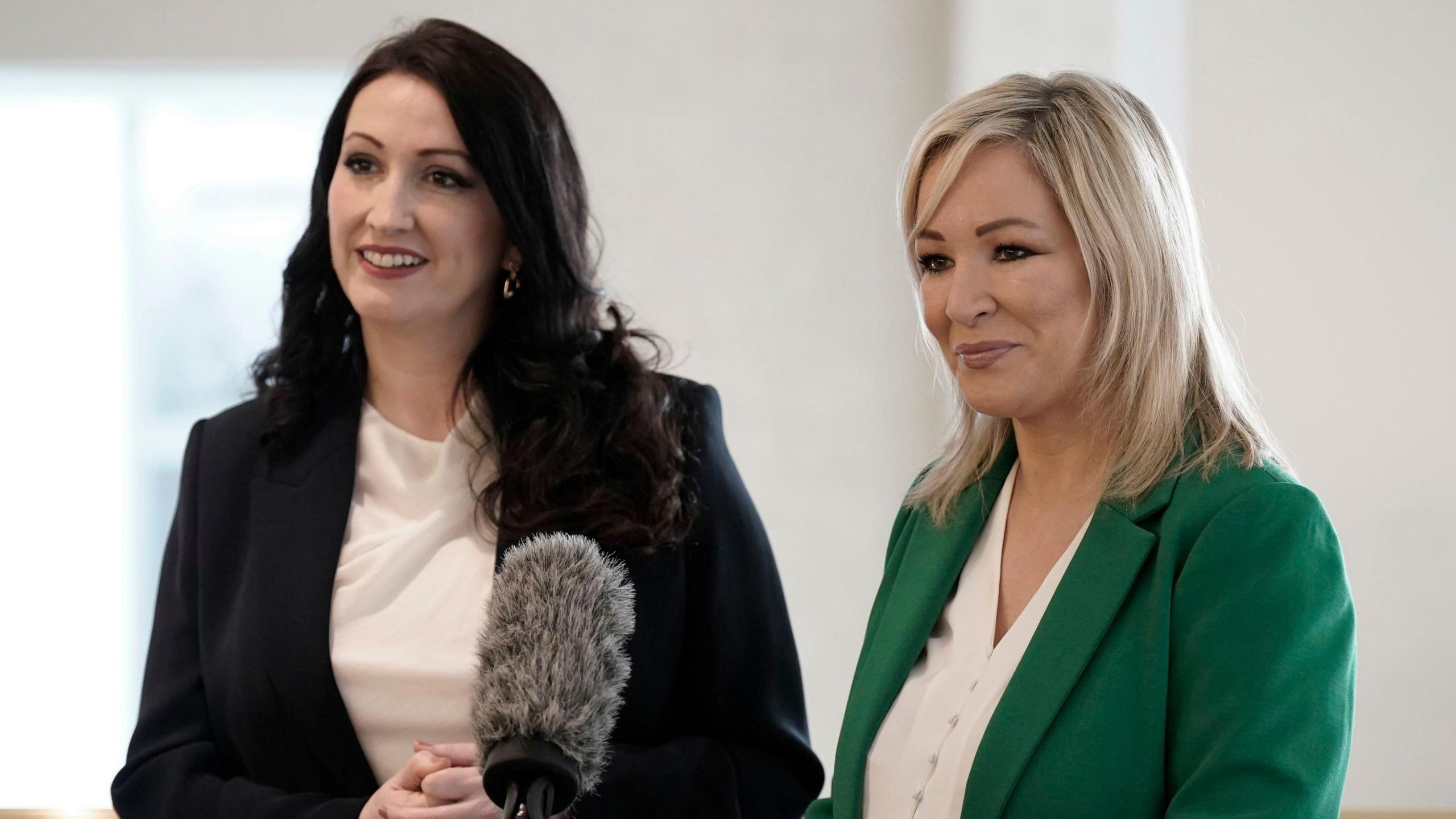 A woman with long brown curly hair is on the left of the image. She is smiling and wearing a black formal blazer and a cream top. The woman on the left has blond shoulder-length hair and is smiling at something off-camera. She is wearing a green formal blazer and a cream top. Between them is a fluffy grey broadcast microphone. 