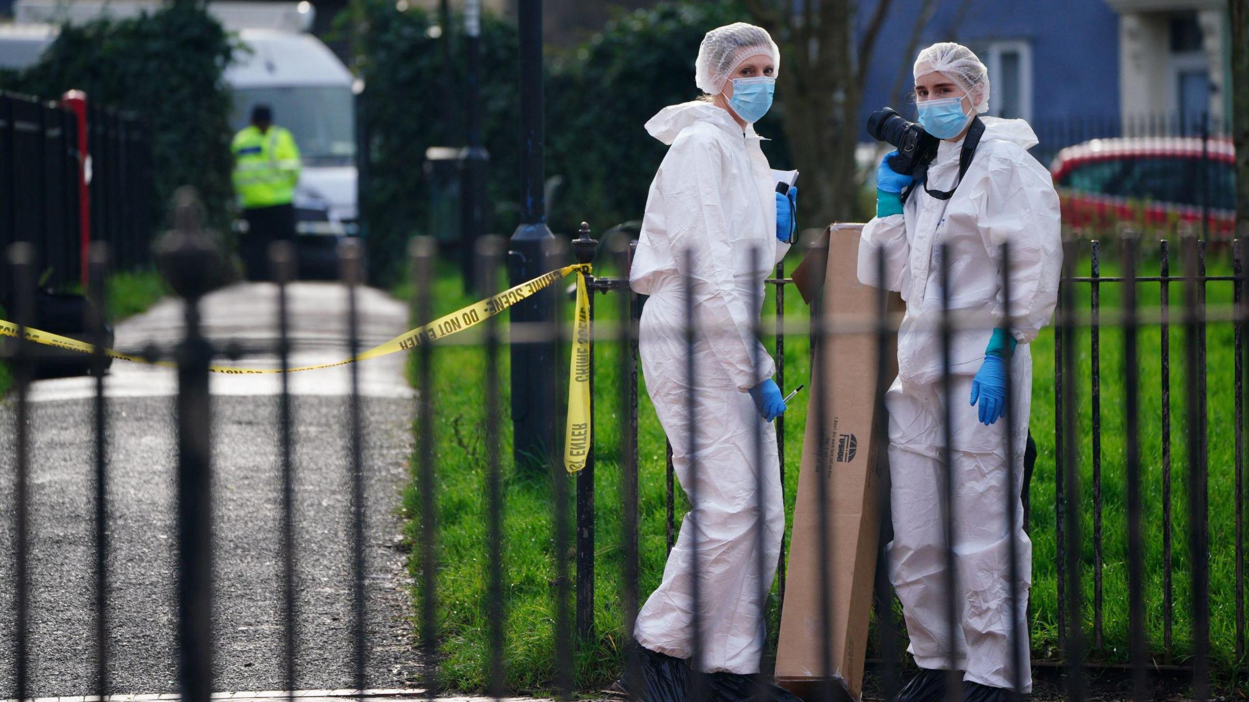 Two police forensic officers in full-length white suits stand in Rawnsley Park in Easton in Bristol. The picture is taken from the other side of a black metal fence