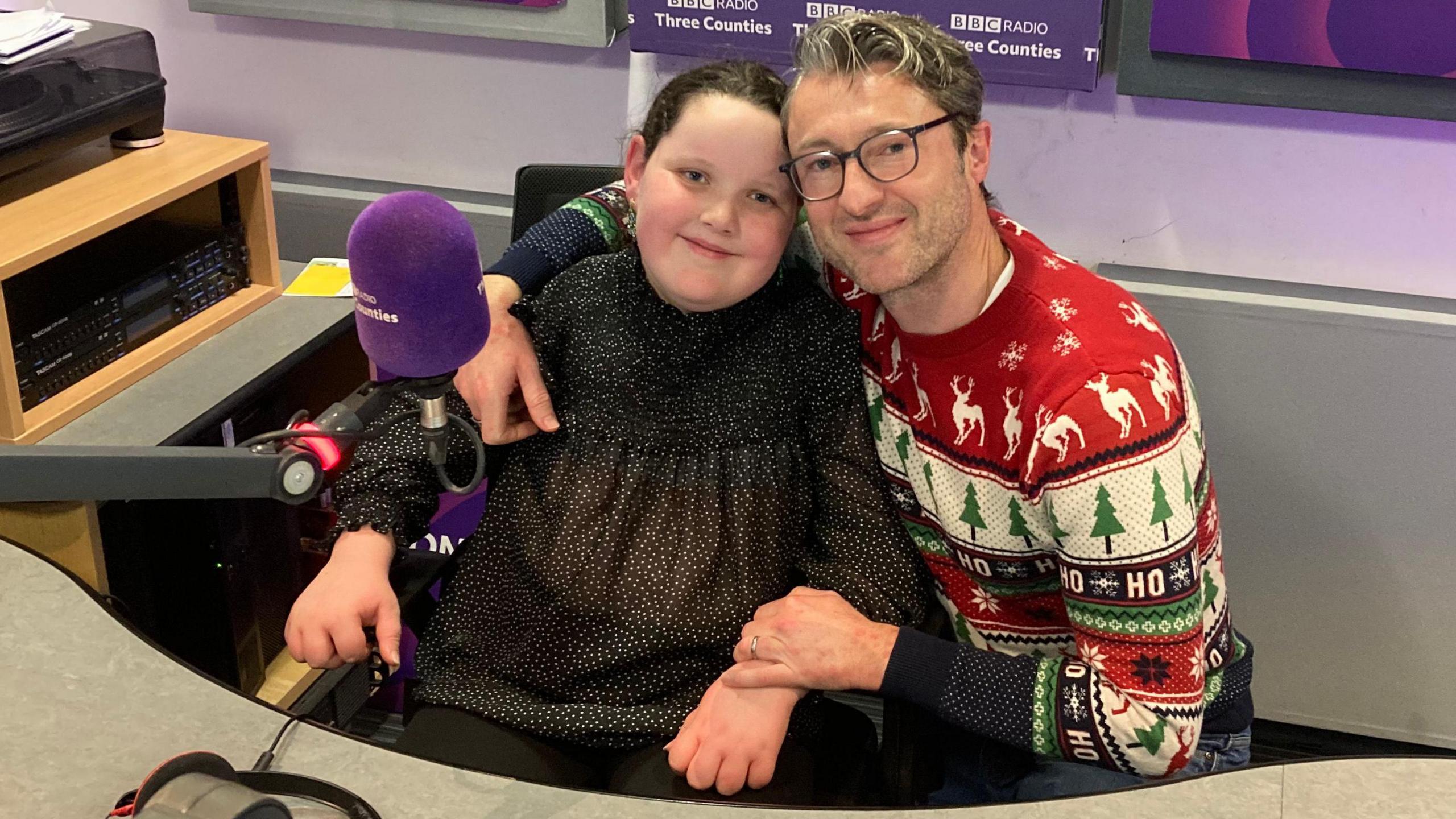 Betsy is sitting in a chair in a radio studio with her father kneeling beside her. They are both smiling at the camera. Betsy is wearing a sparkly black top and her father is wearing spectacles and a Christmas jumper with reindeer and Christmas trees on it