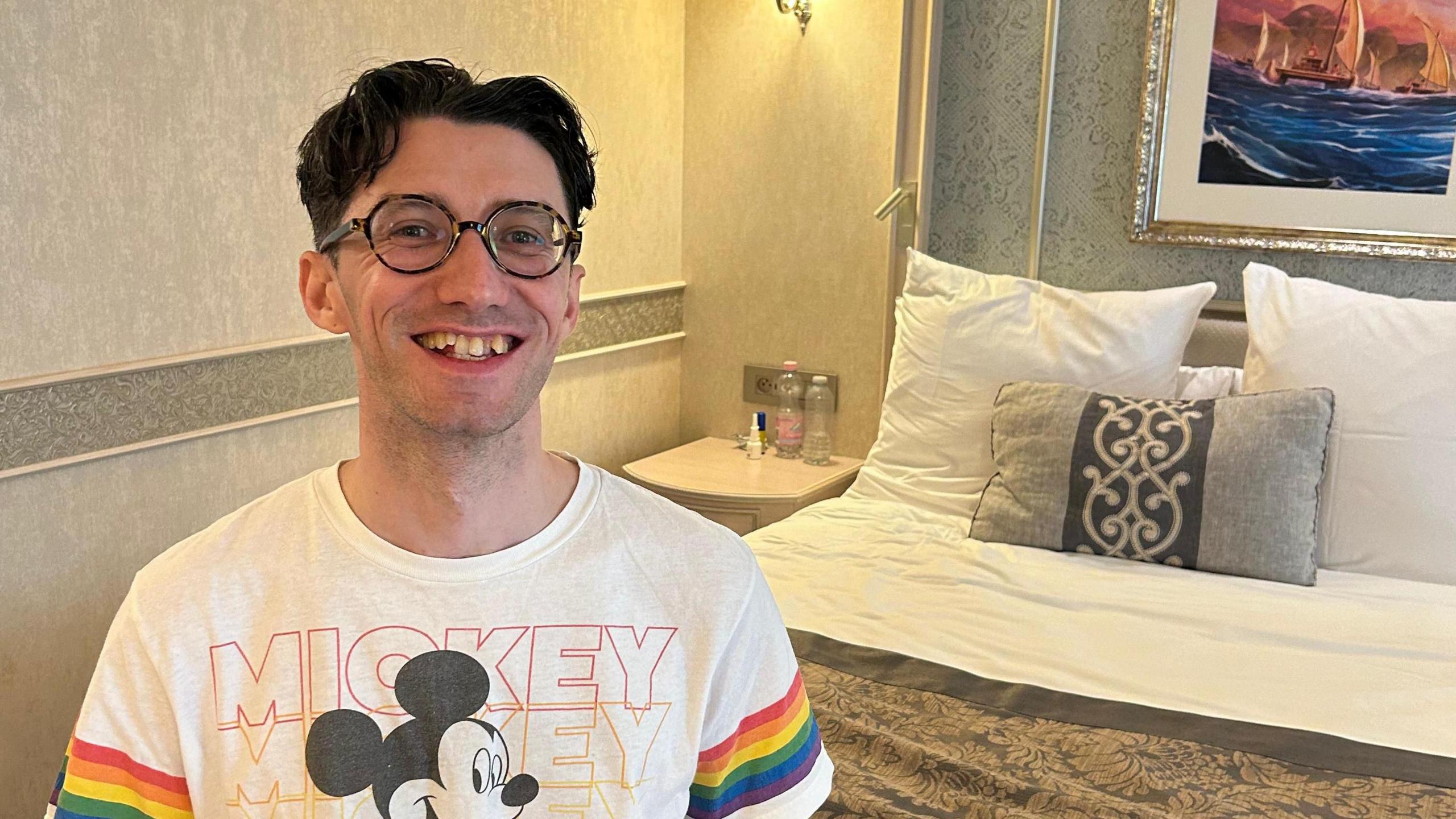 A man wearing round tortoise shell glasses smiles at the camera, perched on the end of a hotel bed. He is wearing a white t-shirt adorned with rainbows on each arm and a picture of Mickey Mouse on the front with the word "Mickey" repeated in different colours in the background.