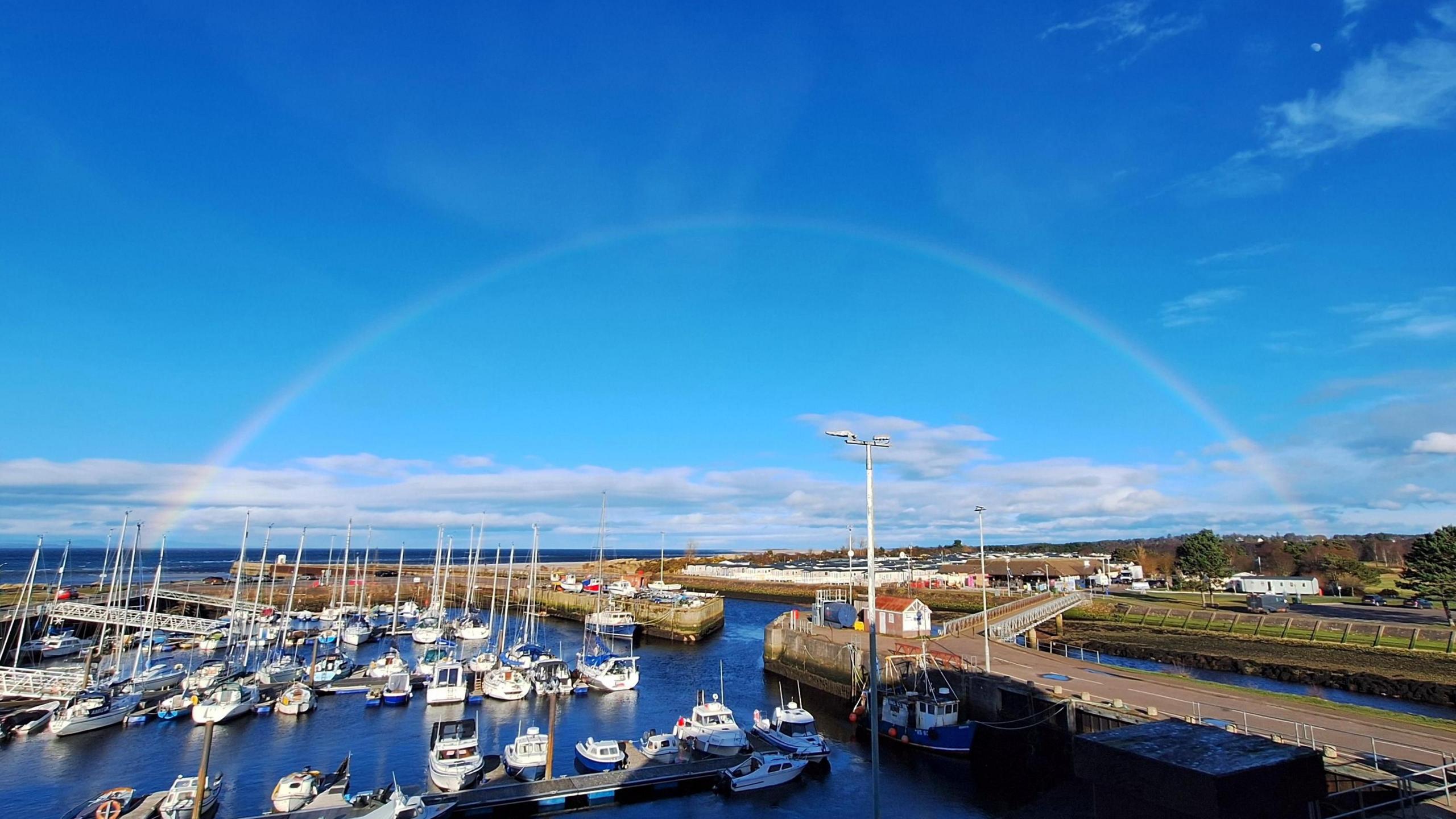 picture overlooking a marina with lots of boats moored up with blue skies and a full rainbow in the sky.