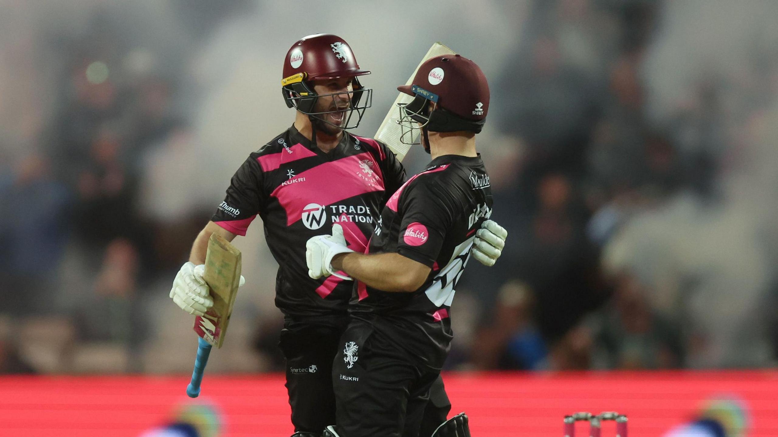 Two Somerset cricketers, wearing the county's purple and black kit, embrace and smile at the end of the T20 Blast final win against Hampshire at Edgbaston. In the background are clouds of white smoke