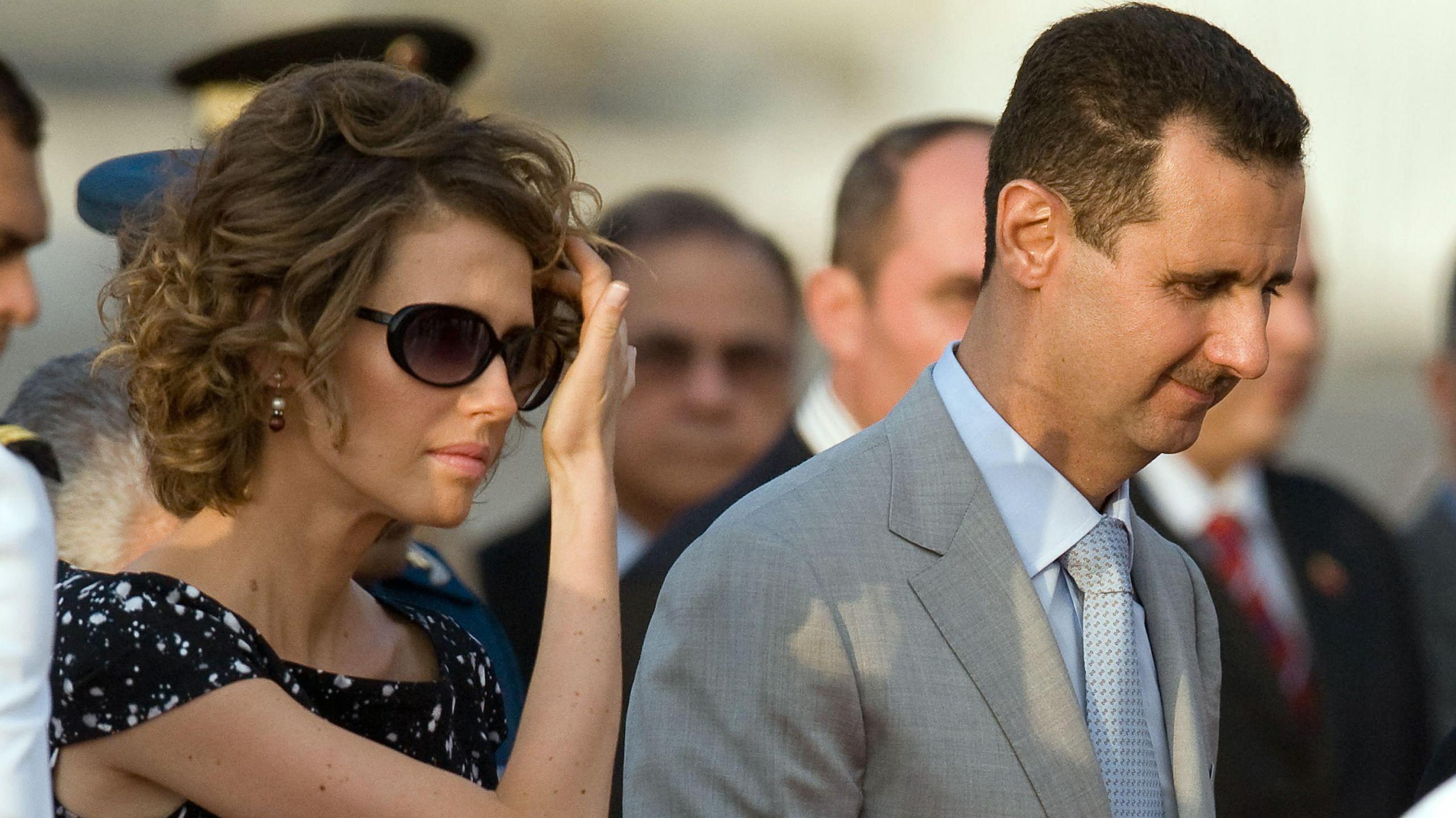 Asma al-Assad, who has short curly hair and is wearing sunglasses and a black outfit, touches her hair. Her husband Bashar is wearing a light grey suit and tie. This is a stock photo from 2010.