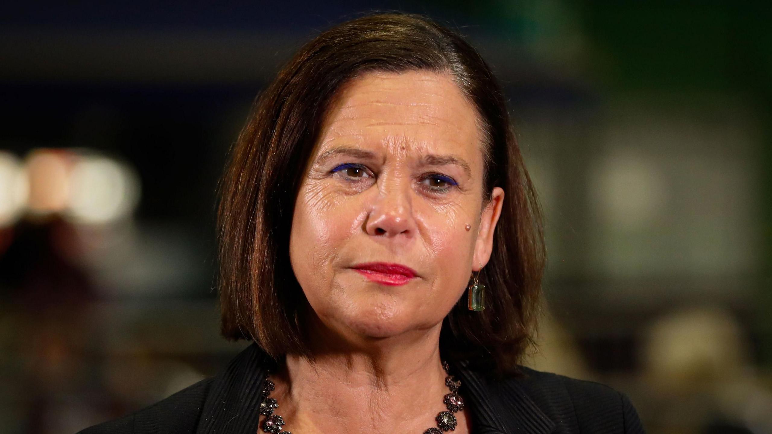 Mary Lou McDonald. She has a brown bob and red lipstick. She wears a pinstripe suit and has a sparkly necklace 