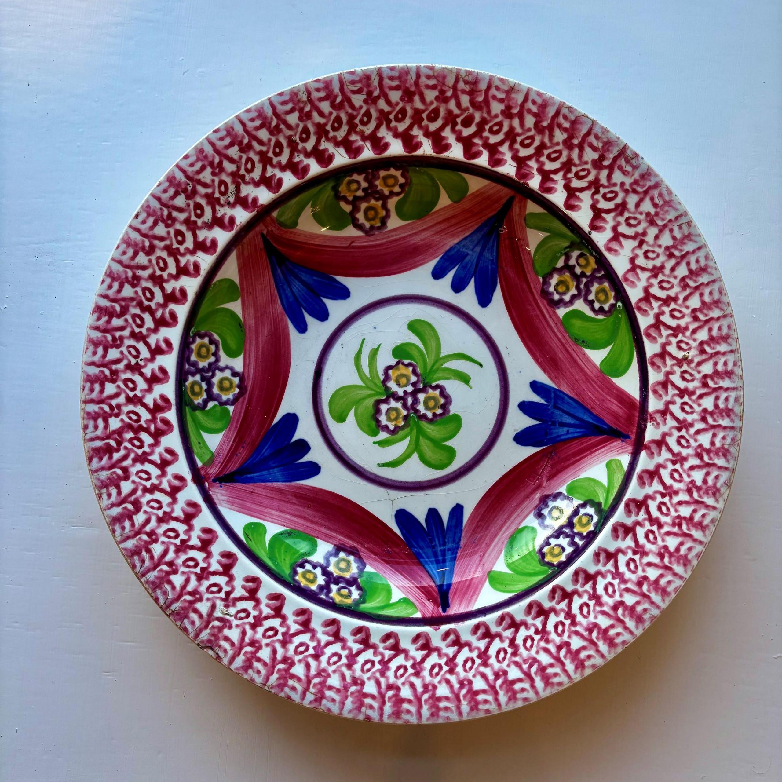 A brightly coloured decorative bowl, with red flowers around the rim of the plate, and a green, red, blue and yellow floral pattern on the inside. Three daisies are painted within a circle in the centre.