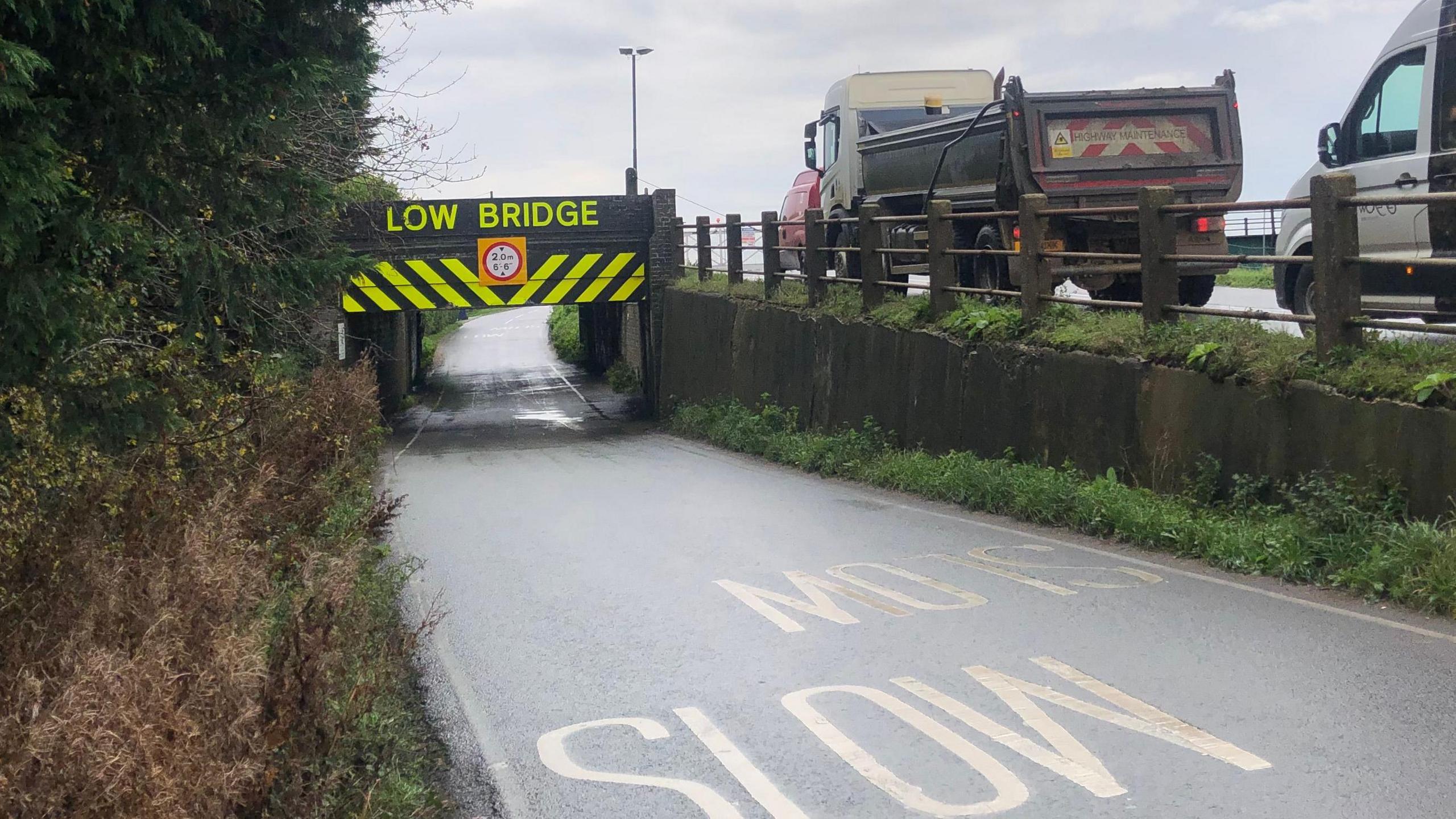 Stonea Road bridge near Manea. It shows two parallel roads, one going under the railway line with slow written in yellow writing on the tarmac. The bridge has yellow signs saying low bridge and yellow warning stripes. Beside it and rising above to the railway line, a flat-bed lorry and a van can be seen waiting to cross the railway line once the barrier is lifted