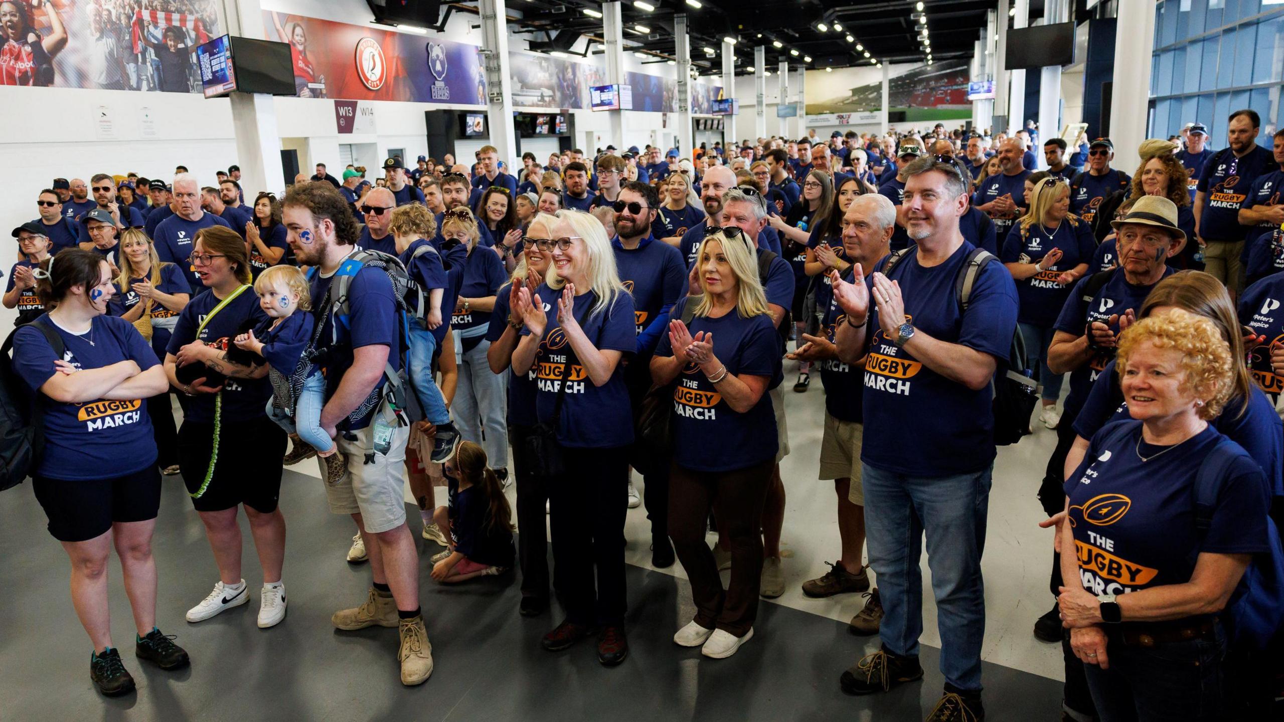 People with the Bristol Bears Foundation in navy the Rugby March t shirts. 
There are a few hundred people in the picture all looking forward and smiling, with many clapping their hands. 