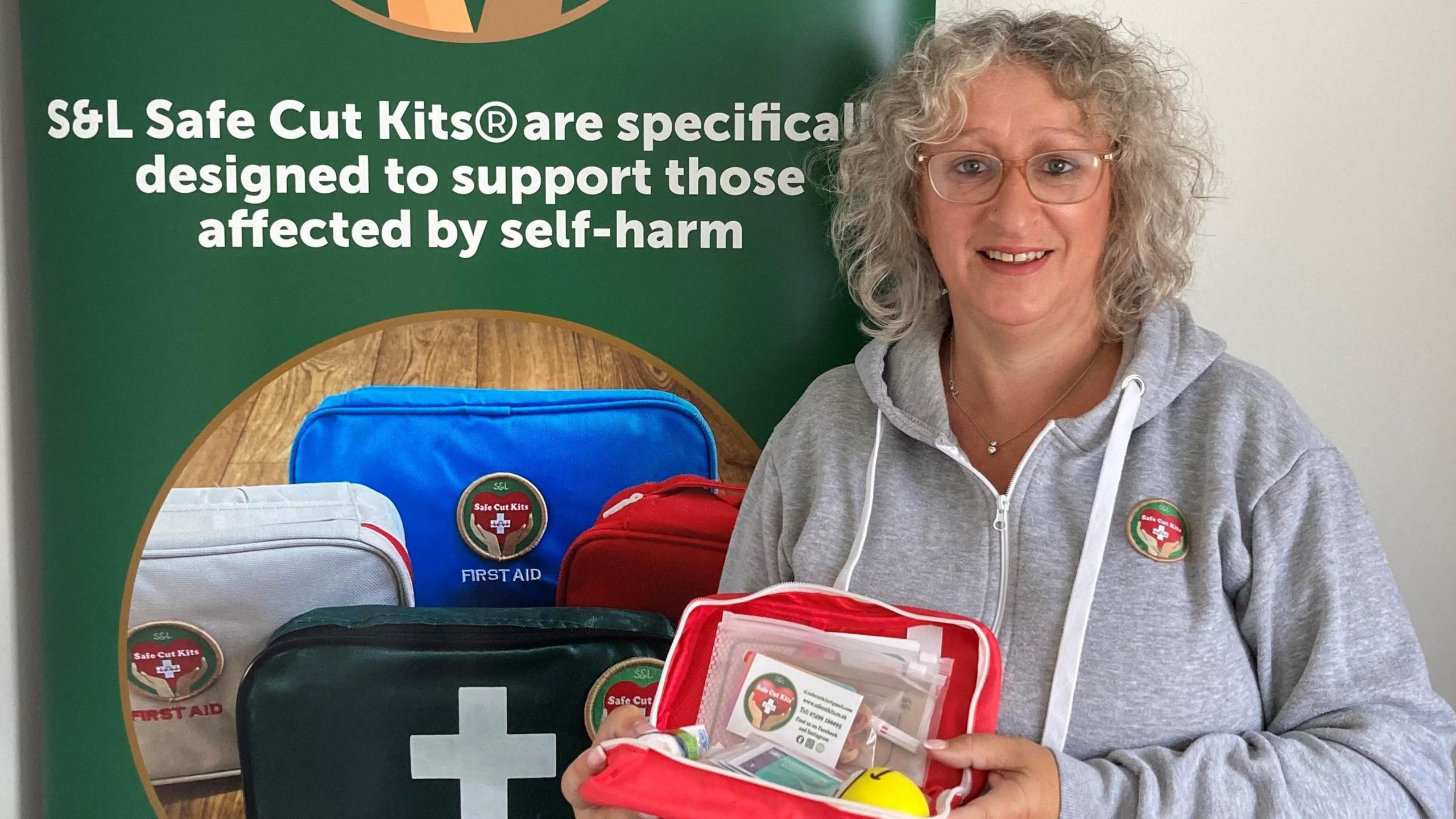 Sally Panks in a grey sweatshirt smiling at the camera. She has curly grey hair and is wearing glasses. She is standing in front of a green poster and is holding a kit. 